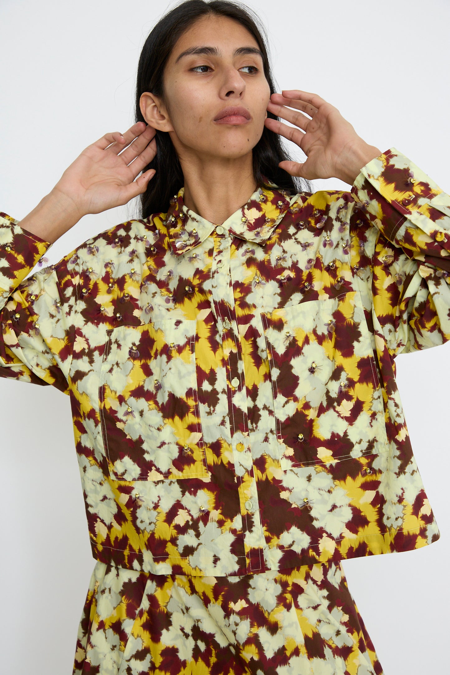 A person wearing the Aria Blouse in Acacia by Ulla Johnson, along with a matching skirt, featuring delicate bead embellishments, is posing with hands near their head.