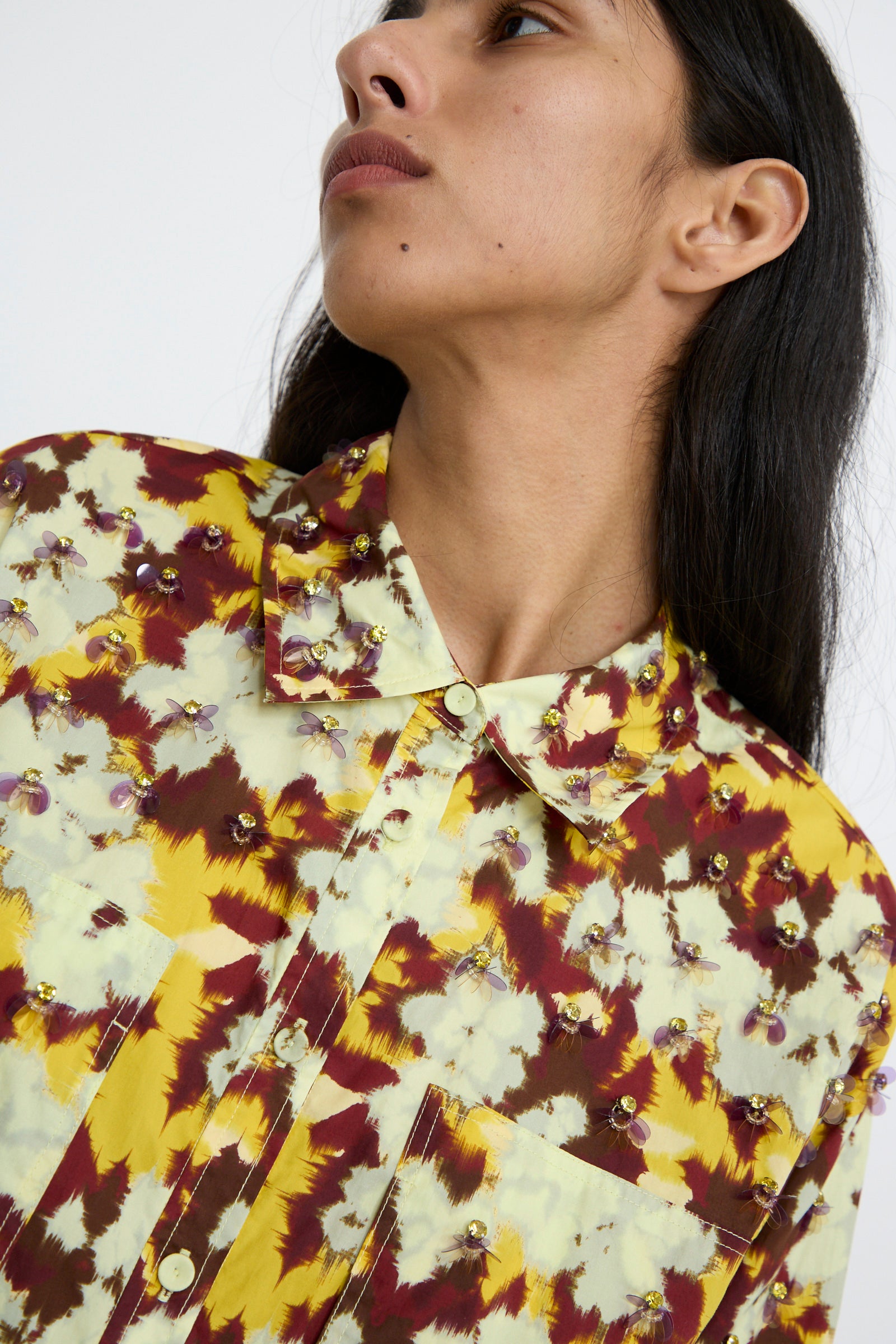 A person wearing the Ulla Johnson Aria Blouse in Acacia, featuring vibrant floral ikat patterns in shades of yellow and maroon with intricate bead embellishments, glances to the side.