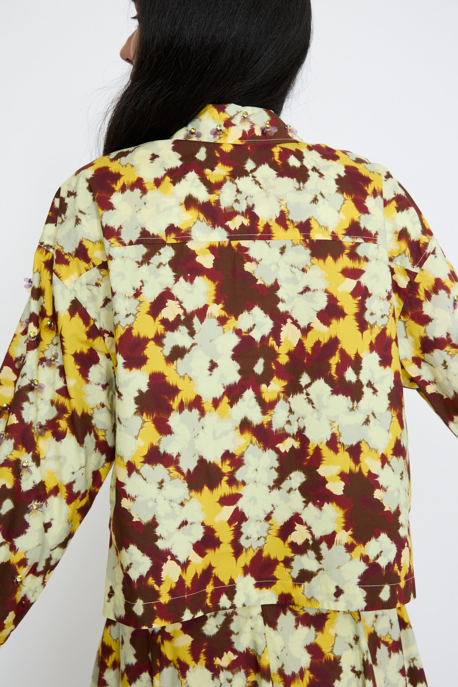 A person is seen from the back, wearing the Aria Blouse in Acacia by Ulla Johnson, featuring a colorful abstract pattern with yellow, white, and maroon hues against a white background. 