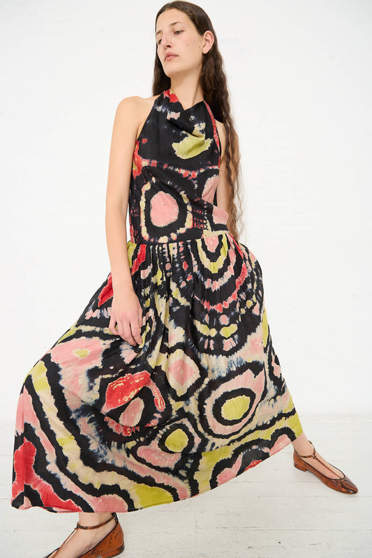 Person standing against a white background, wearing the Assa Dress in Mirage by Ulla Johnson—a colorful, patterned silk dress with a halter top and full skirt featuring a unique motif—paired with brown shoes. Hair is long and loose.
