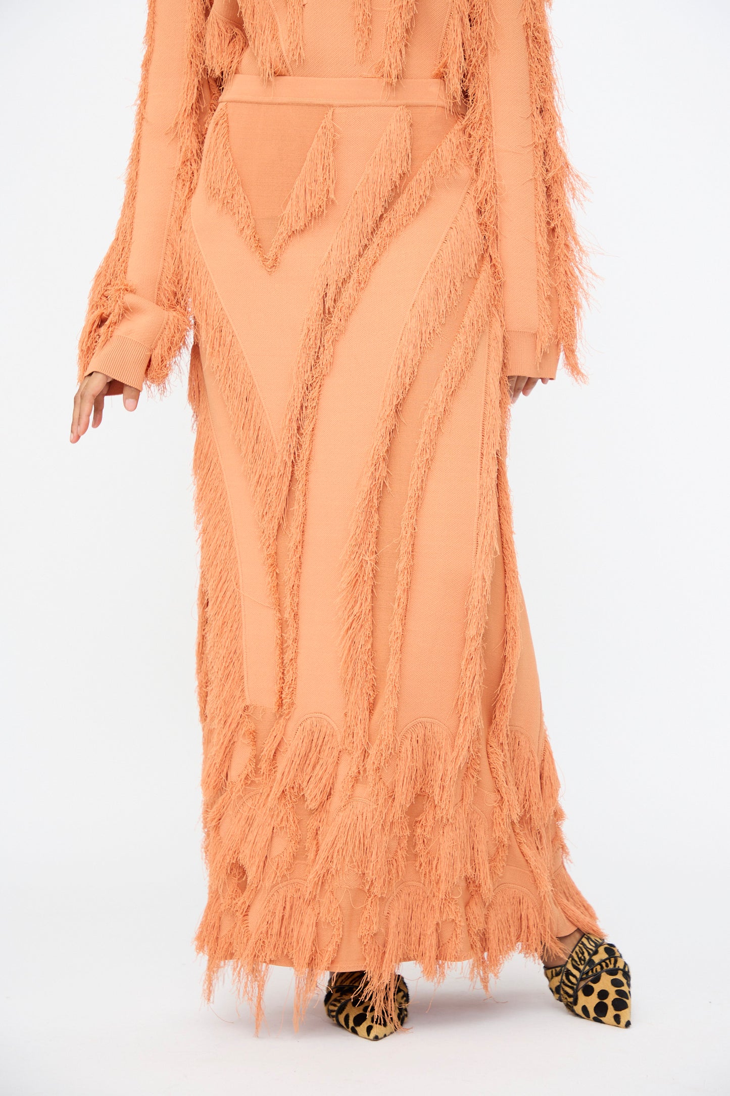 A person wears the Ulla Johnson Astrid Maxi Skirt in Tulip, featuring peach jacquard fabric, fringe details, and an elastic waist, paired with leopard print shoes, standing against a plain white background.