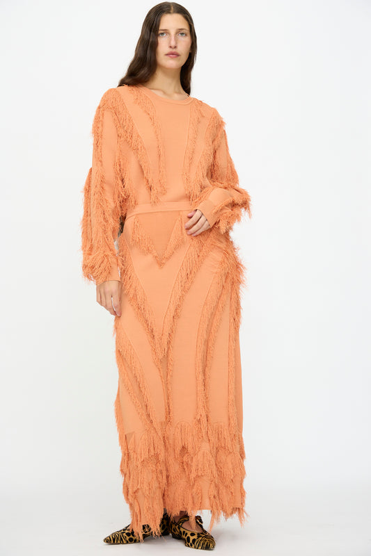 A person stands against a white background wearing Ulla Johnson’s Astrid Maxi Skirt in Tulip, paired with a matching peach long-sleeve top and animal print shoes, the skirt featuring jacquard texture and fringe detailing.