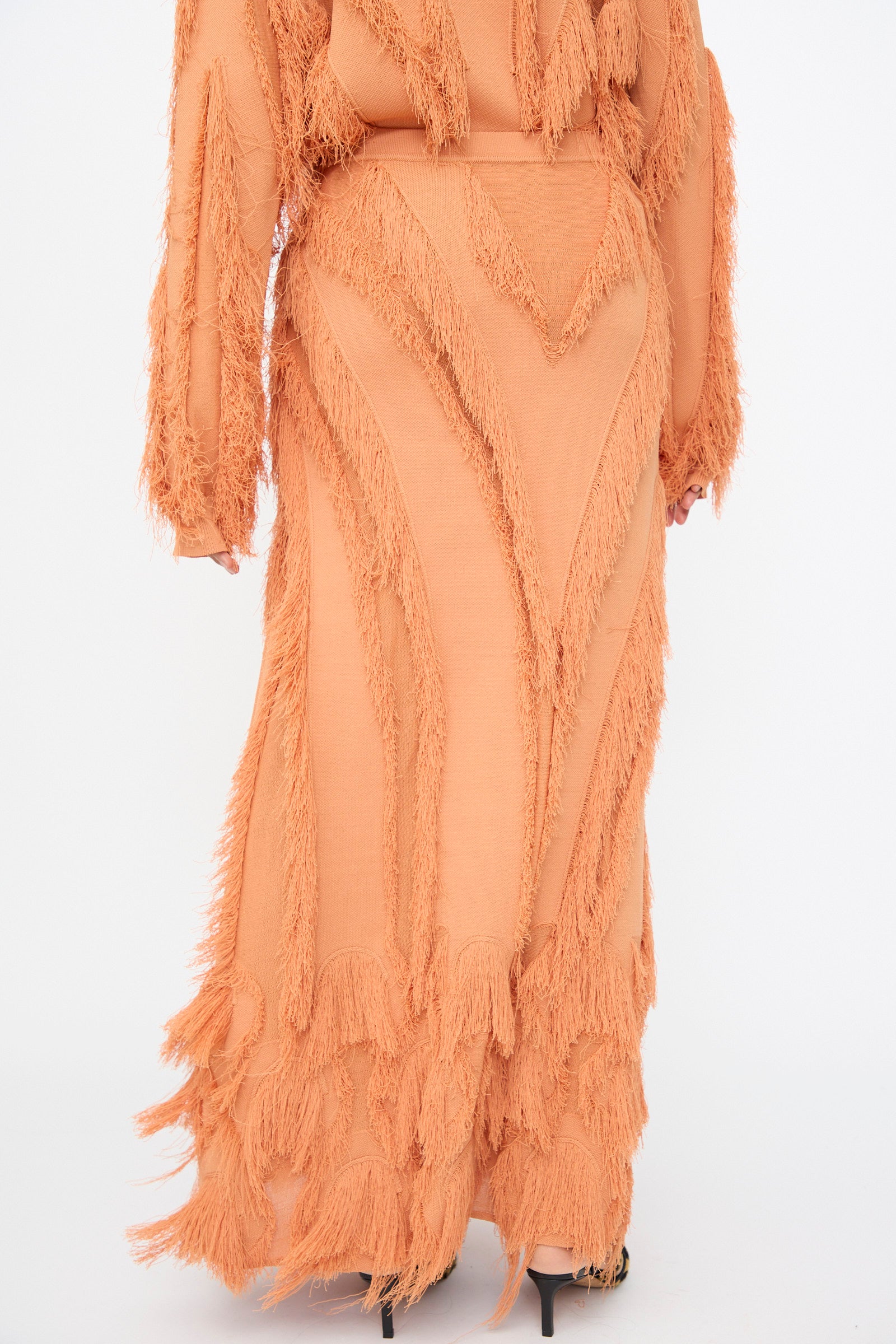 A person wearing the Astrid Maxi Skirt in Tulip by Ulla Johnson, a long peach jacquard skirt with textured fringe and patterns, stands against a plain white background.