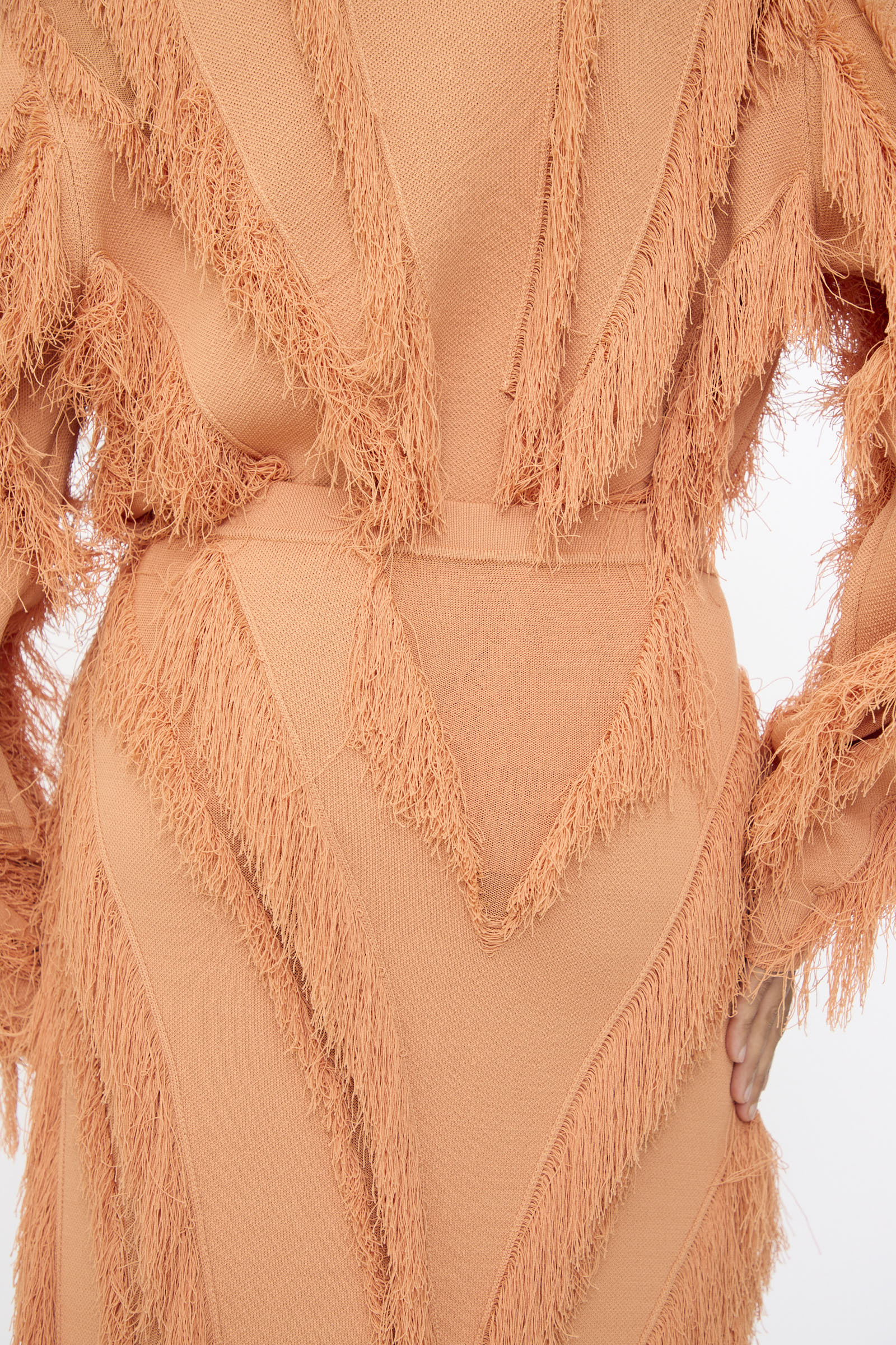 A person models the Ulla Johnson Astrid Maxi Skirt in Tulip, a textured peach jacquard skirt with fringed zigzag patterns, shown from the back with one hand on their hip.