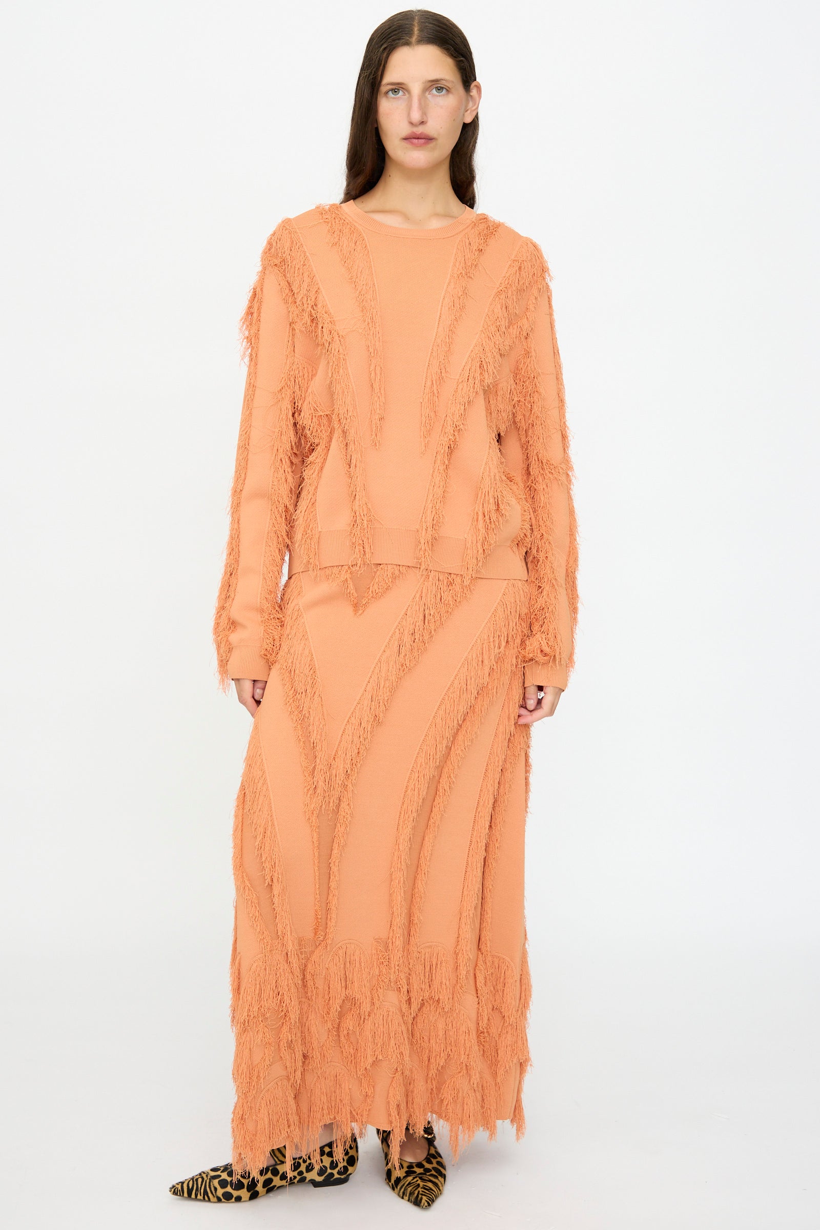 A person stands facing forward wearing the Ulla Johnson Aubree Knit Pullover in Tulip and a matching long skirt with fringe pattern details, paired with leopard print shoes, against a plain white background.