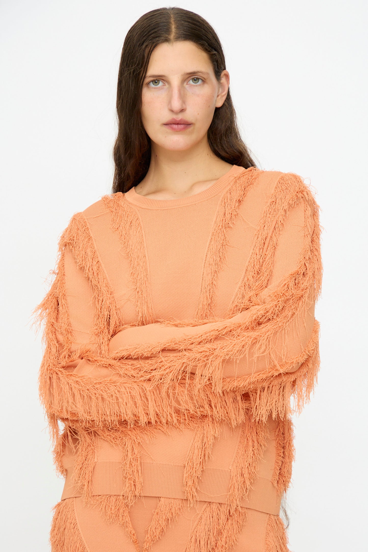 A woman with long brown hair wears the Ulla Johnson Aubree Knit Pullover in Tulip, featuring fringe detailing, while standing against a plain white background.