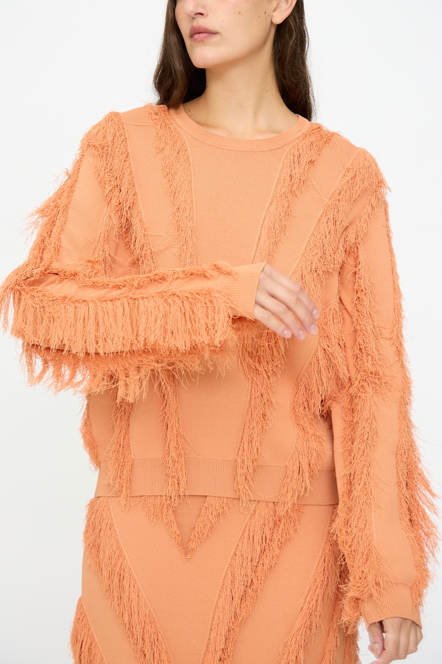 A woman wears the Ulla Johnson Aubree Knit Pullover in Tulip—a peach jacquard sweater with fringed details and matching skirt—standing against a plain white background.