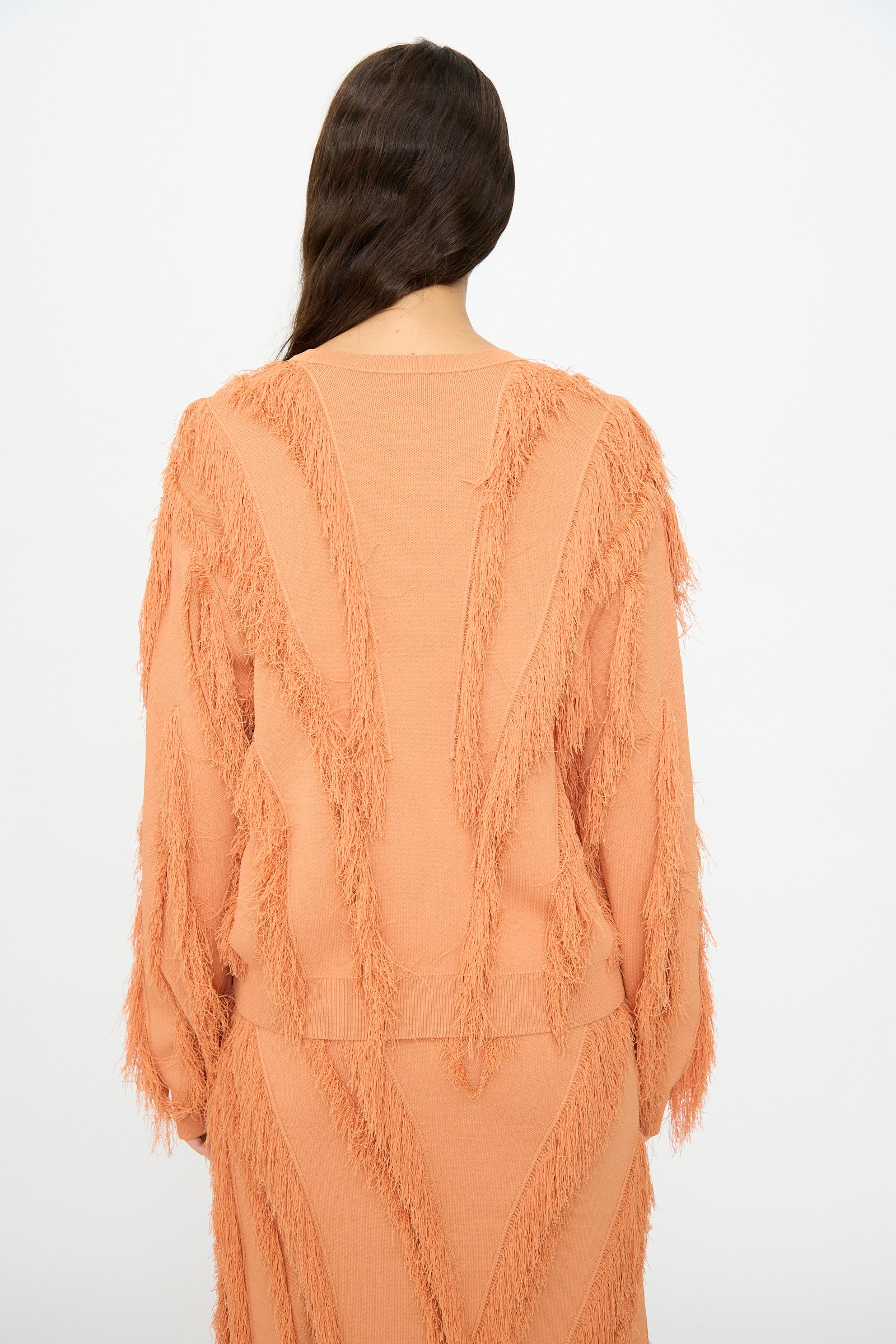 A person with long brown hair, shown from the back, wears the Ulla Johnson Aubree Knit Pullover in Tulip—a peach jacquard sweater with textured fringe on the sleeves, back, and skirt.