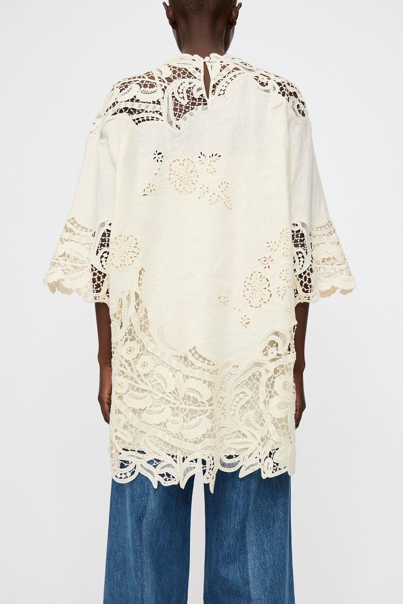 A person is seen from the back wearing Ulla Johnson's Botanical Linen Lace Brinna Top in Pristine—an oversized, white tunic with floral lace embroidery and scalloped edges—paired with blue jeans against a plain background.