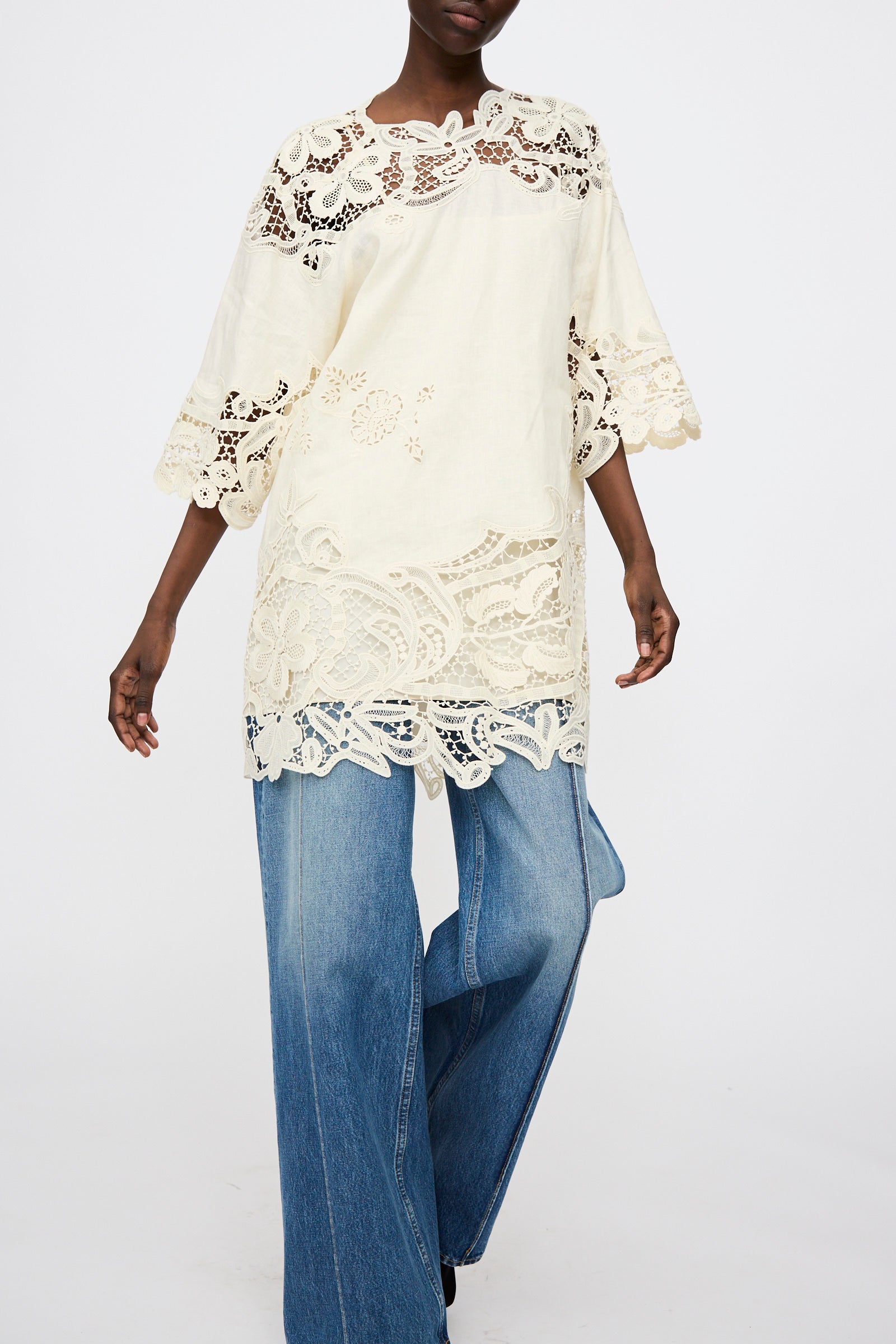 A person wears the Ulla Johnson Botanical Linen Lace Brinna Top in Pristine, featuring flared sleeves and a keyhole fastening, paired with wide-leg blue jeans against a plain background.
