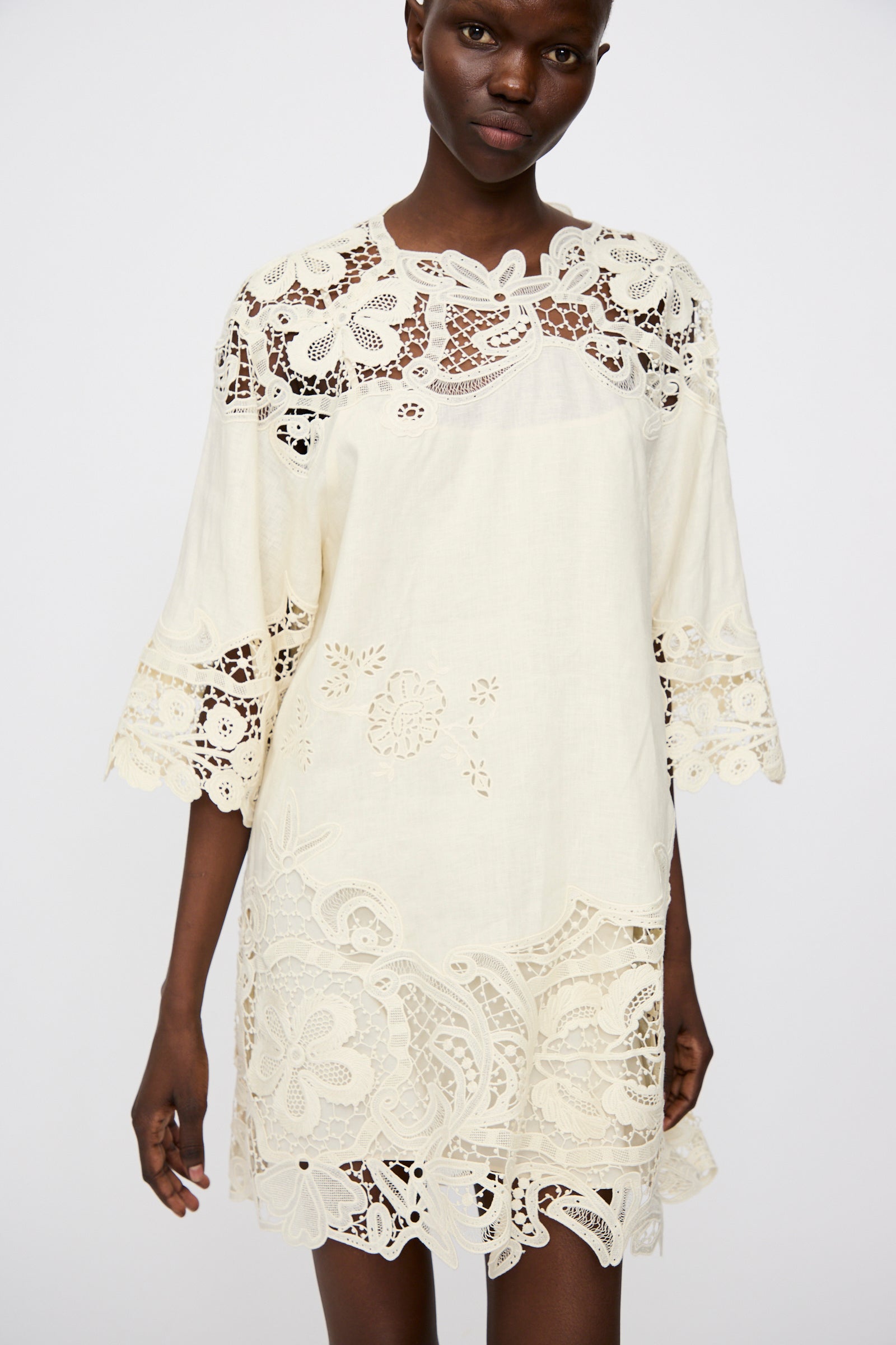 Someone wearing the Ulla Johnson Botanical Linen Lace Brinna Top in Pristine, a loose-fitting cream tunic adorned with intricate floral lace on the sleeves, hem, and upper chest.