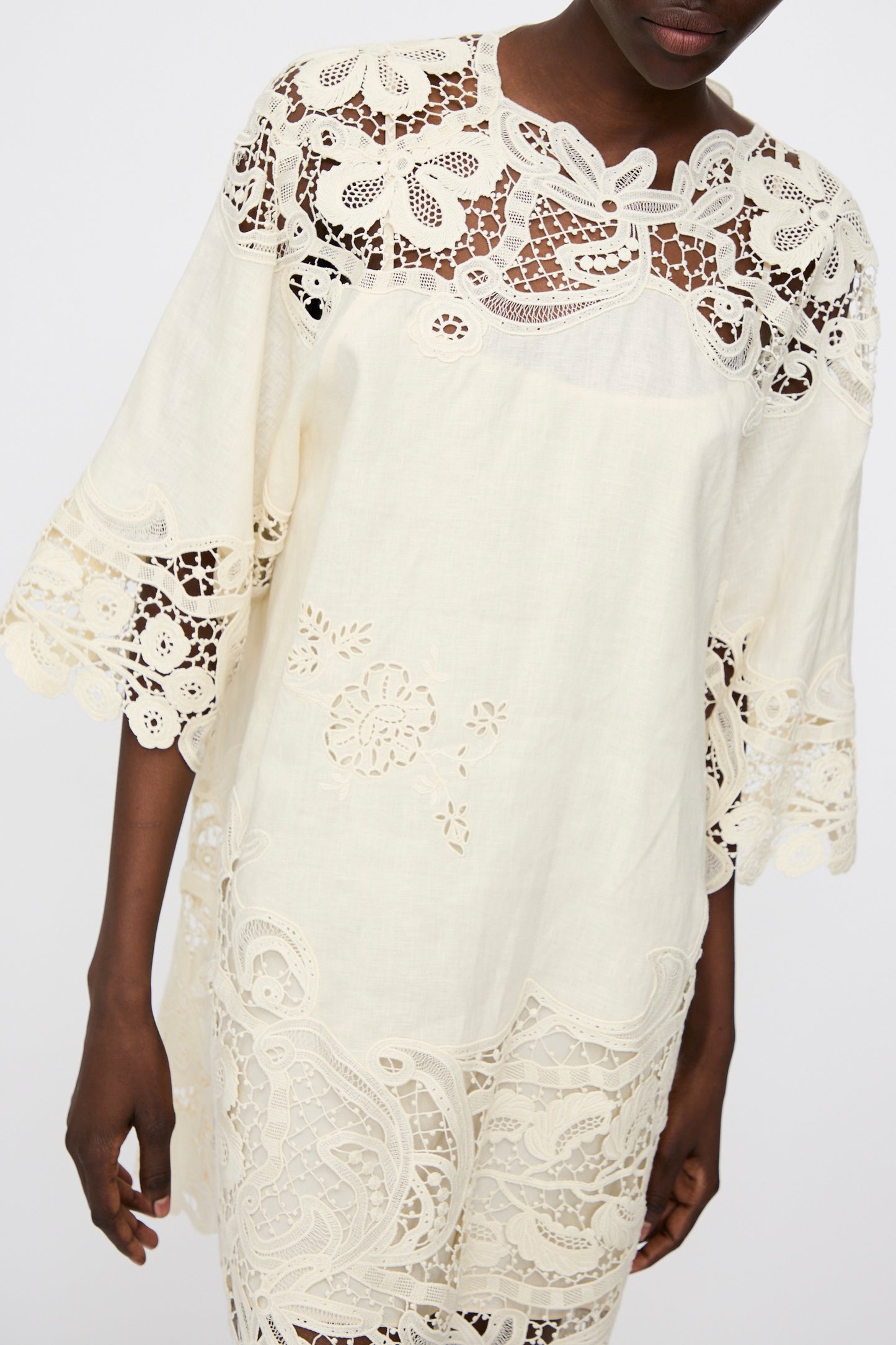 A person wearing the Ulla Johnson Botanical Linen Lace Brinna Top in Pristine, featuring a loose fit with intricate floral lace and embroidered cutouts on the sleeves, bodice, and hem.