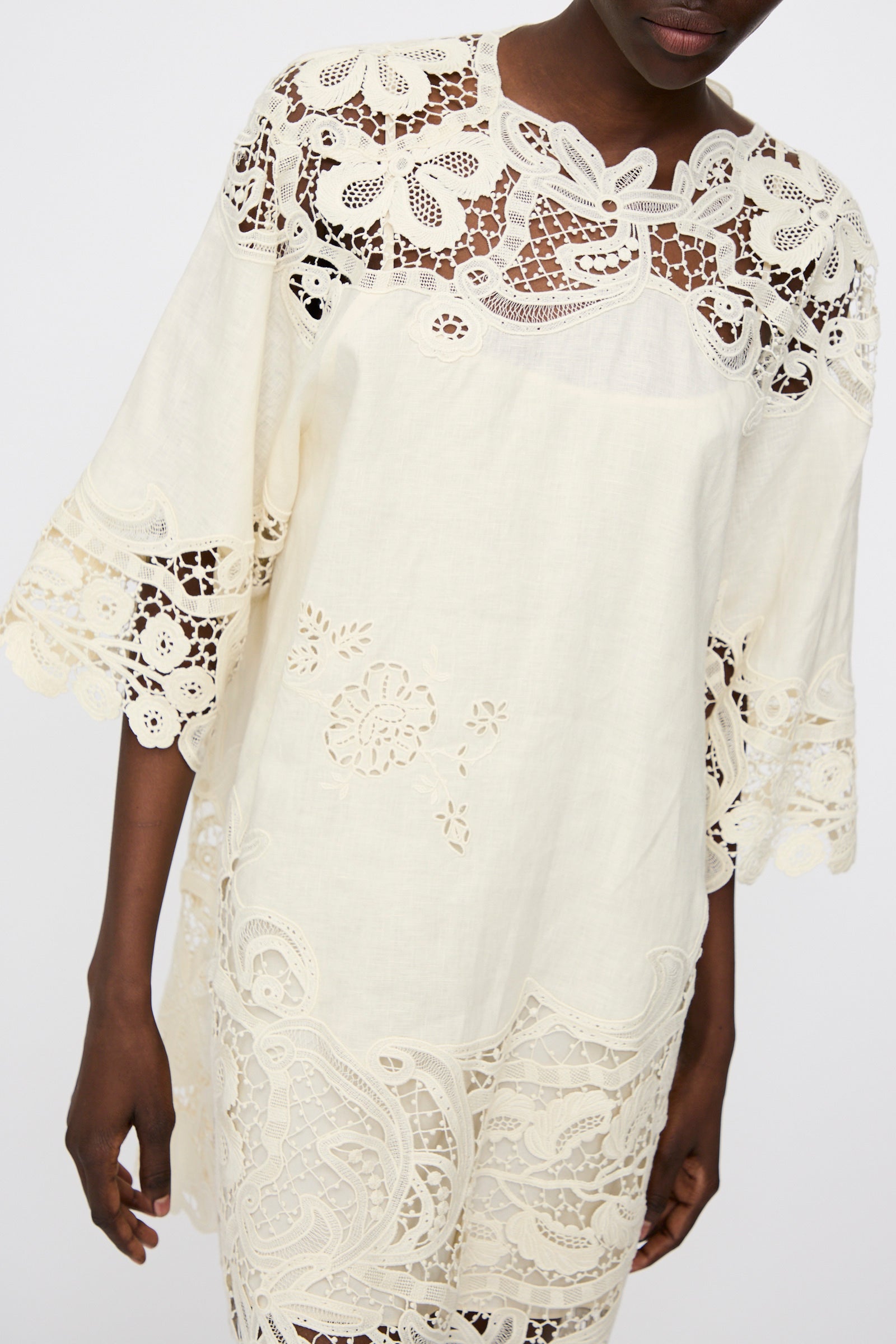 A person wearing the Ulla Johnson Botanical Linen Lace Brinna Top in Pristine, featuring a loose fit with intricate floral lace and embroidered cutouts on the sleeves, bodice, and hem.