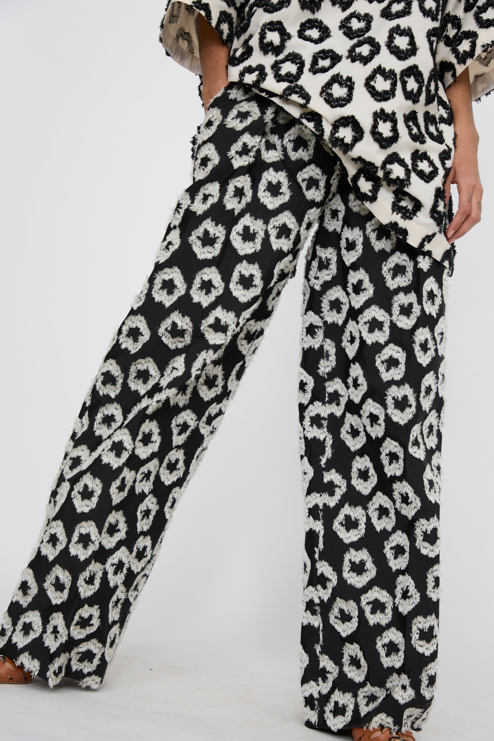 A person is wearing the Cai Pant in Eclipse by Ulla Johnson, featuring a black and white abstract design with wide legs, with one hand in their pocket against a white background. 