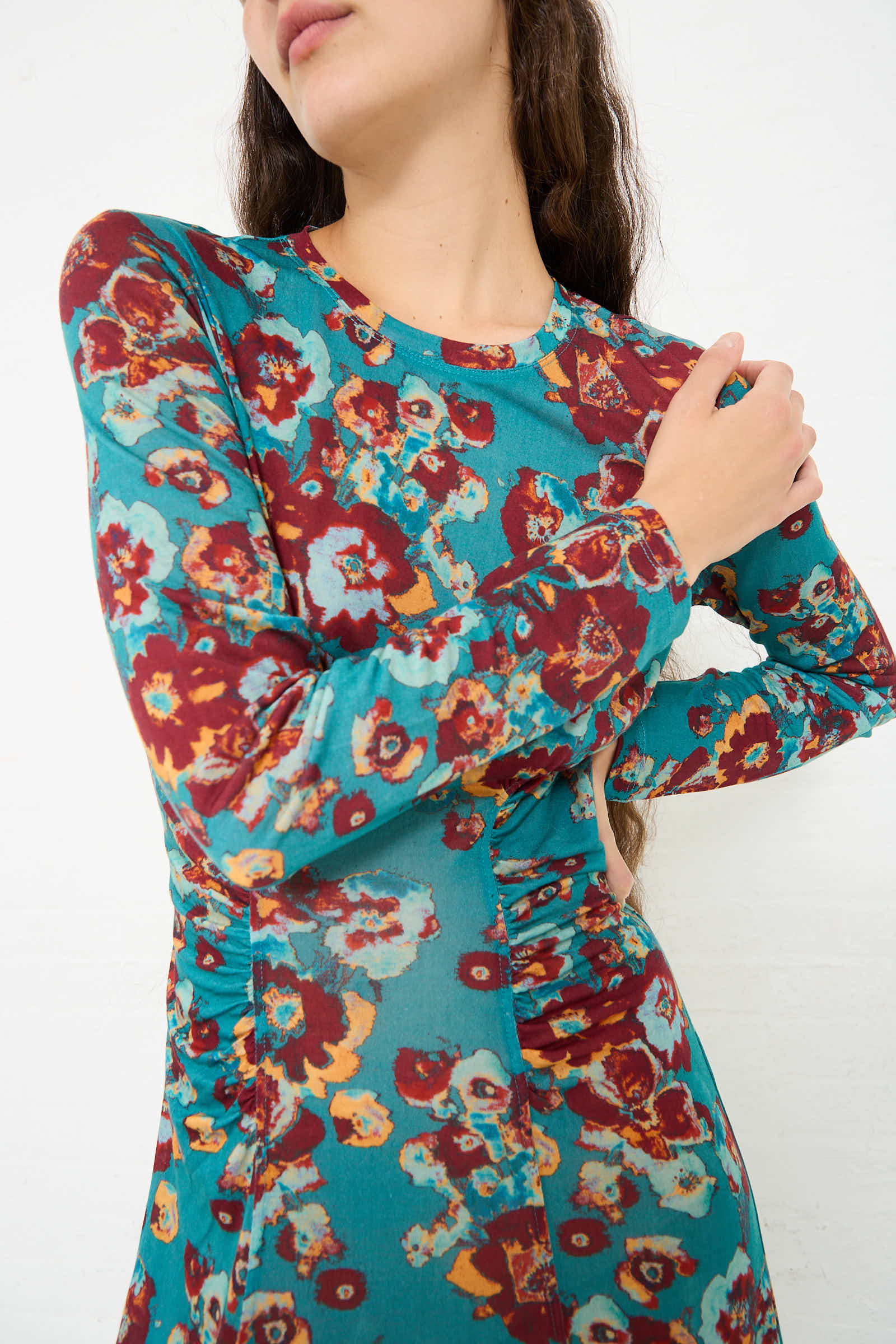 A person standing with one arm crossed, wearing a stylish Ulla Johnson Ceryse Dress in Jade Floral featuring a bold floral print in shades of turquoise, red, yellow, and white. The person's head is partially out of the frame.