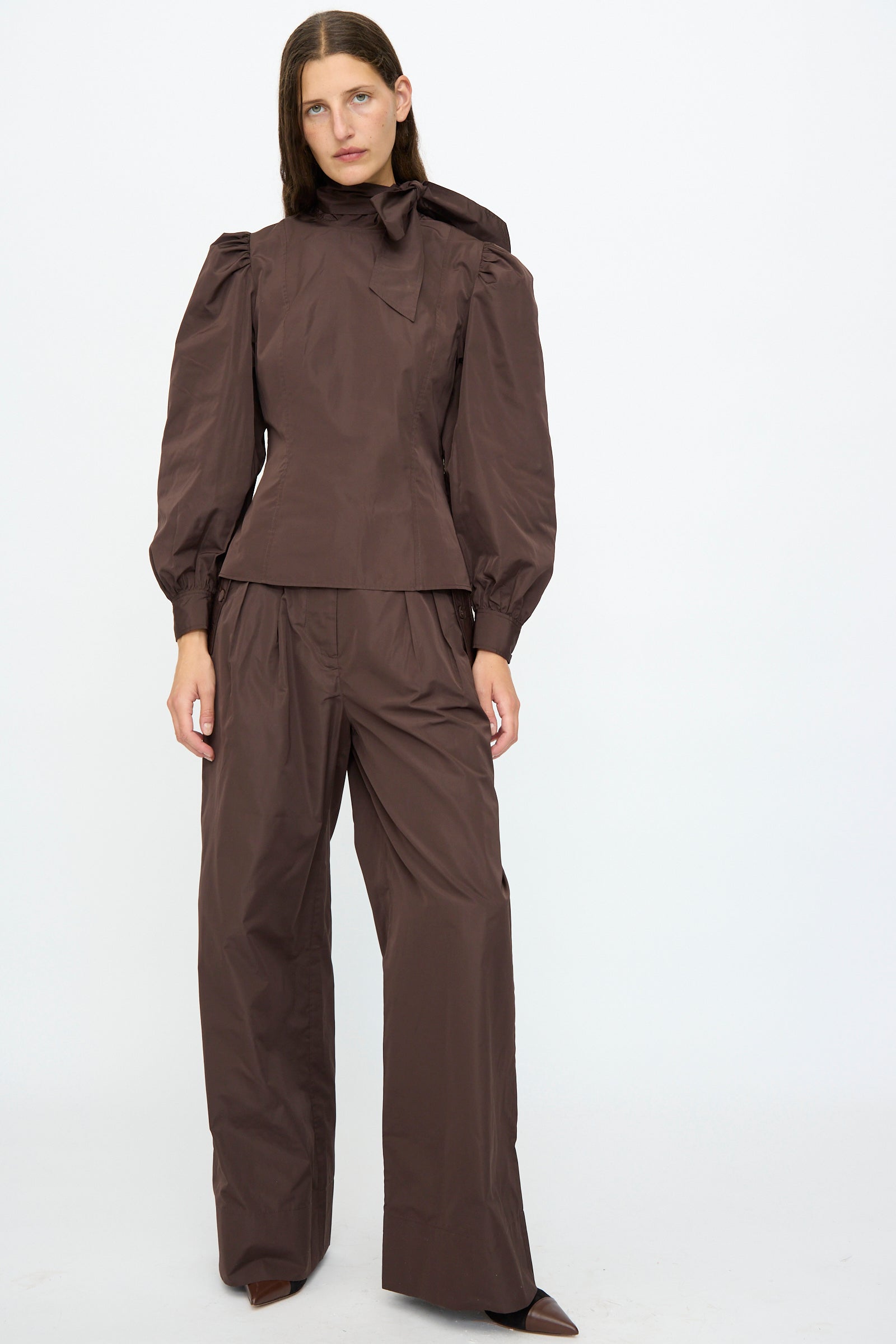 A person wearing Ulla Johnson's Charlie Silk Pant in Sequoia with a brown long-sleeve blouse featuring puffed shoulders and a bow collar, stands against a plain white background.
