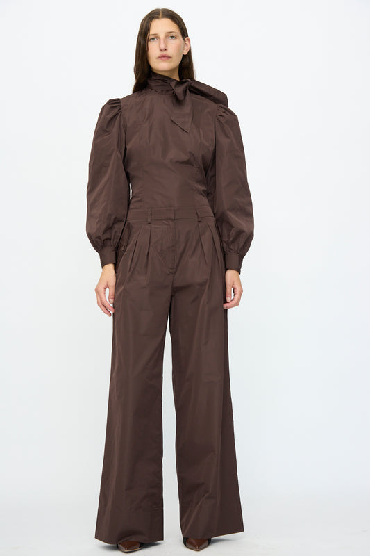 A person stands against a plain background, wearing Ulla Johnson's Charlie Silk Pant in Sequoia, featuring a taffeta finish, puffed shoulders-inspired style, wide legs, and refined high-neck detailing.