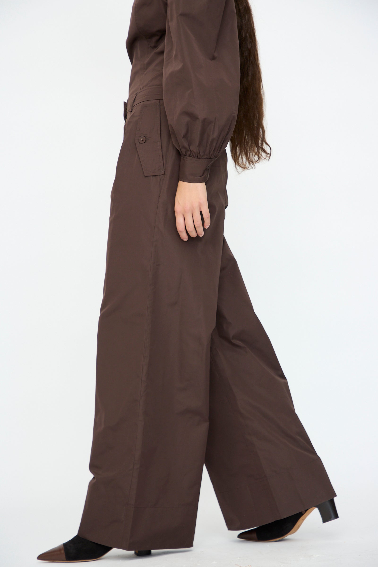 Model shown from the side in Ulla Johnson's Charlie Silk Pant in Sequoia, a brown high-rise, wide-leg style, paired with black pointed-toe shoes against a plain background.