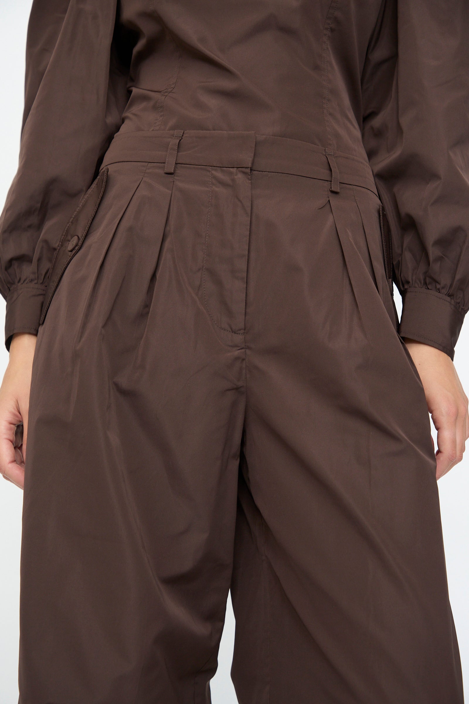 Close-up of a person wearing Ulla Johnson’s Charlie Silk Pant in Sequoia, high-waisted with wide legs and pleats, paired with a matching brown top, both crafted from taffeta-finished habotai.