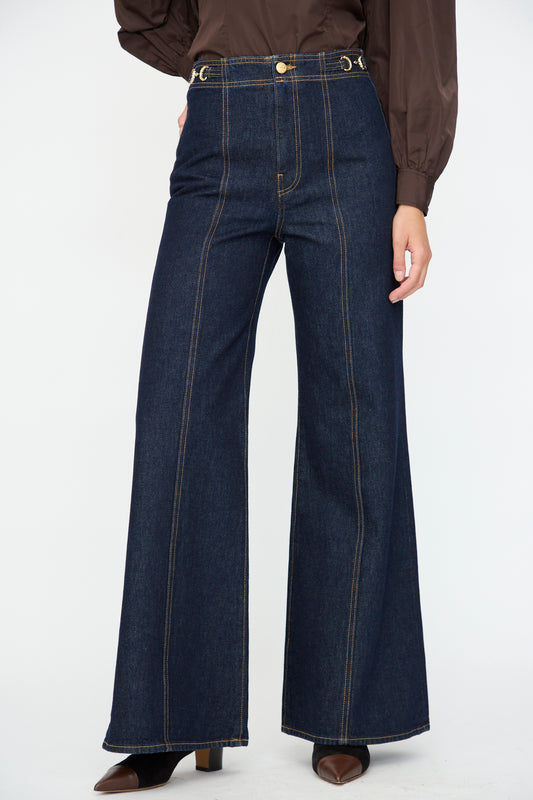 Wearing the Ulla Johnson Delphine High Rise Jean in Tigris, featuring deep indigo cotton with flared legs and visible stitching, styled with a brown long-sleeve top and heeled shoes.