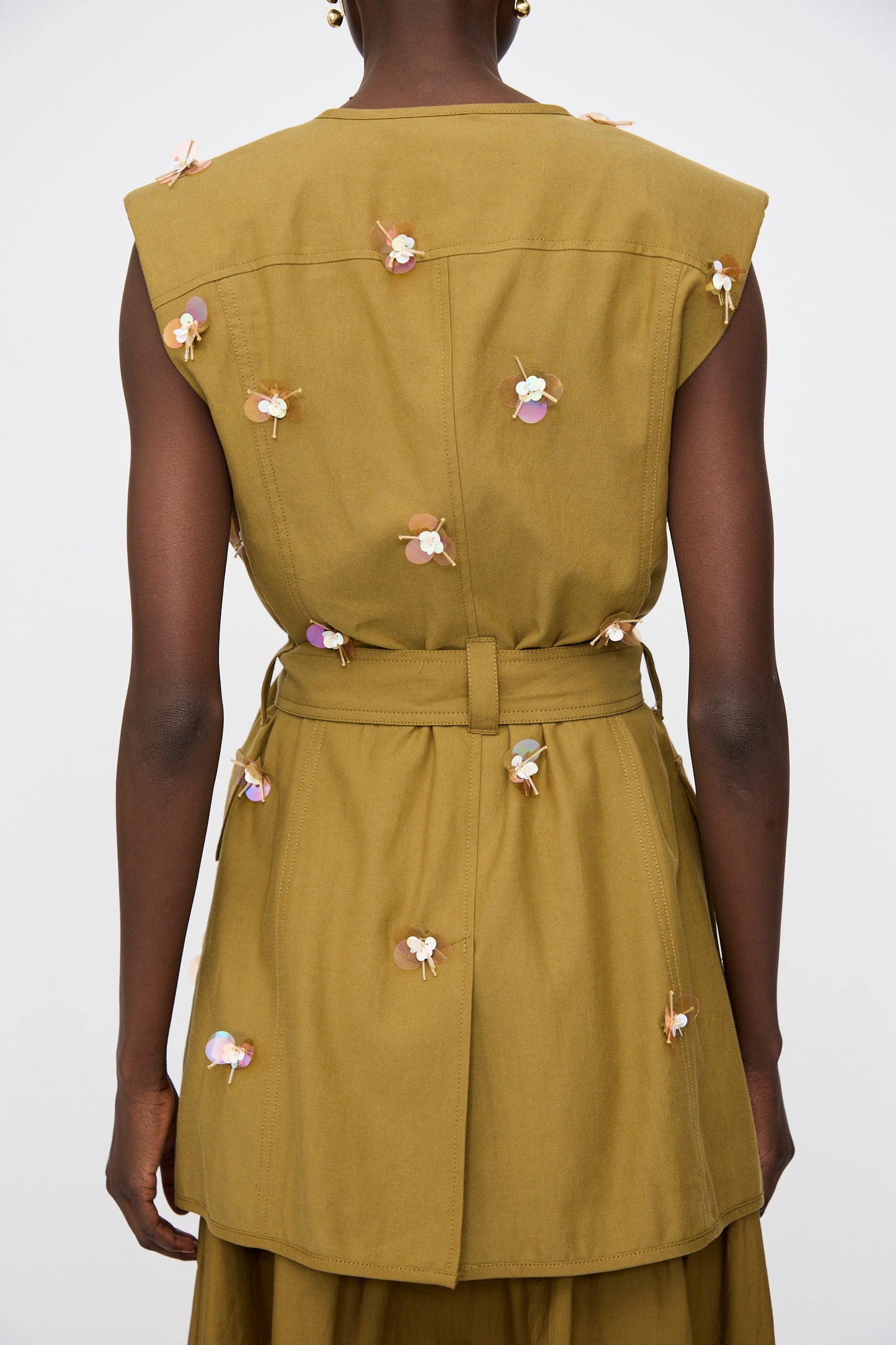 A person shown from the back wears the Ulla Johnson Embroidered Cotton Twyla Vest in Lichen—a sleeveless, tailored piece belted at the waist and adorned with small iridescent floral paillettes.