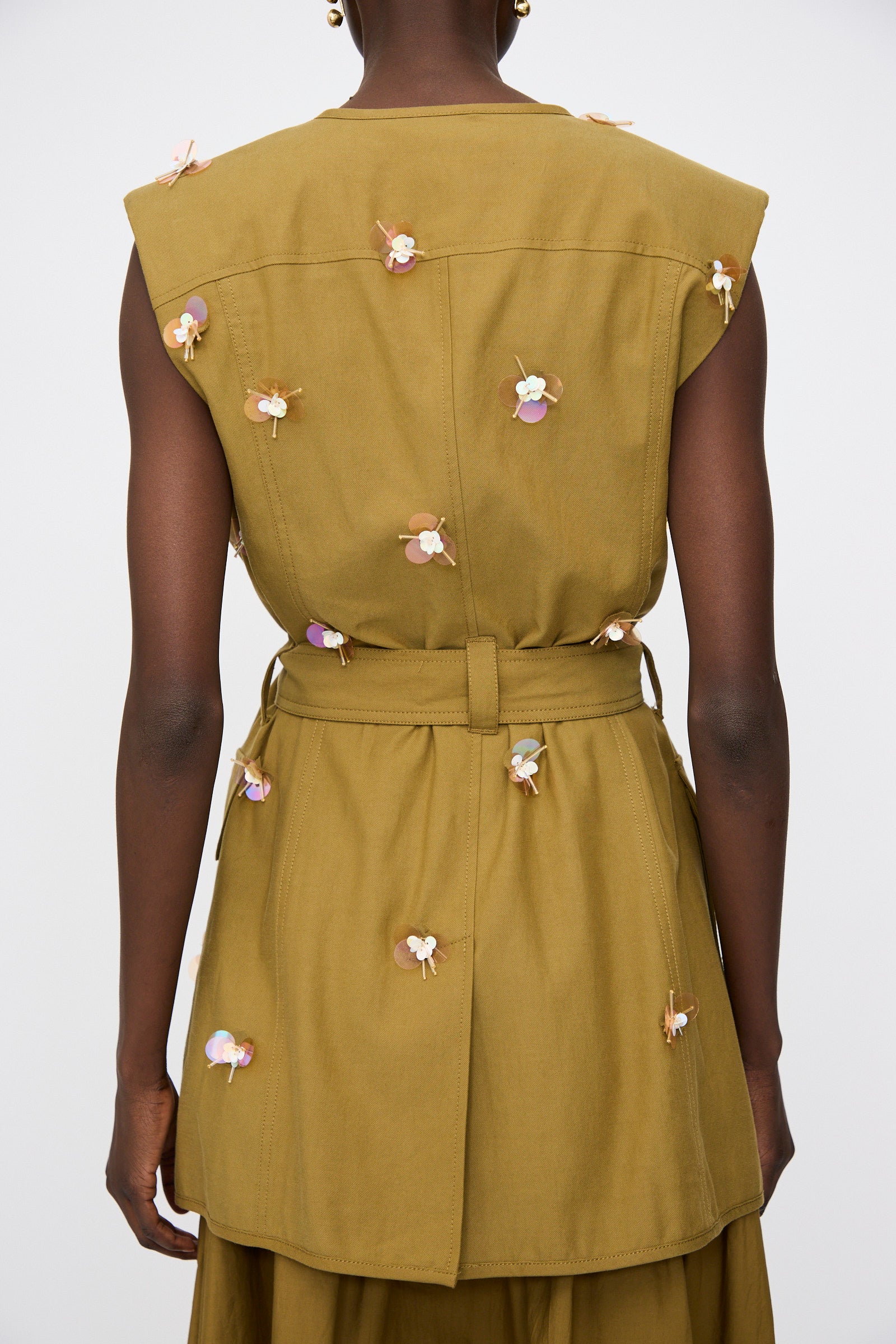 A person shown from the back wears the Ulla Johnson Embroidered Cotton Twyla Vest in Lichen—a sleeveless, tailored piece belted at the waist and adorned with small iridescent floral paillettes.