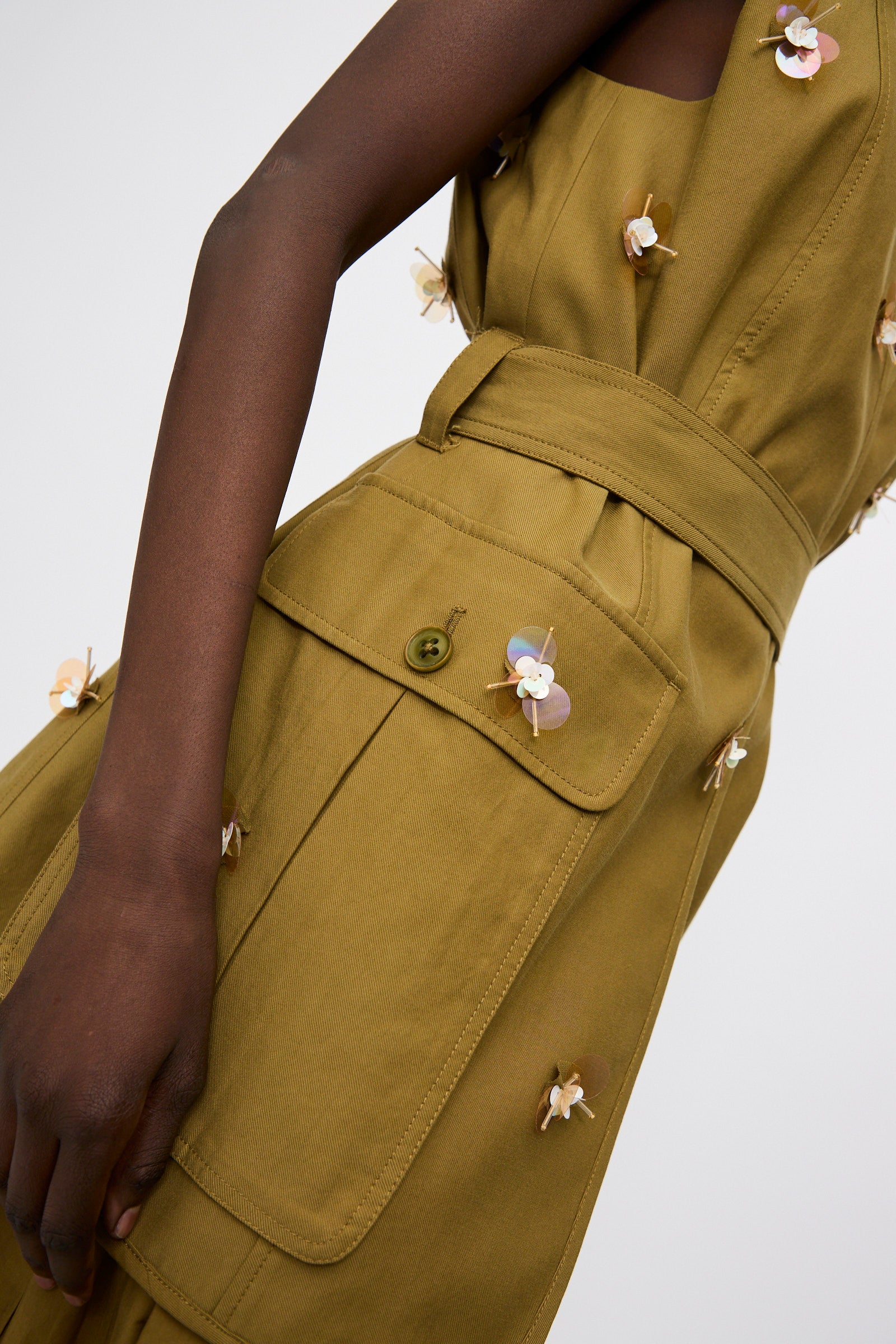 A person wearing the Ulla Johnson Embroidered Cotton Twyla Vest in Lichen, a tailored olive-green sleeveless dress featuring large pockets and delicate 3D paillette floral embellishments.