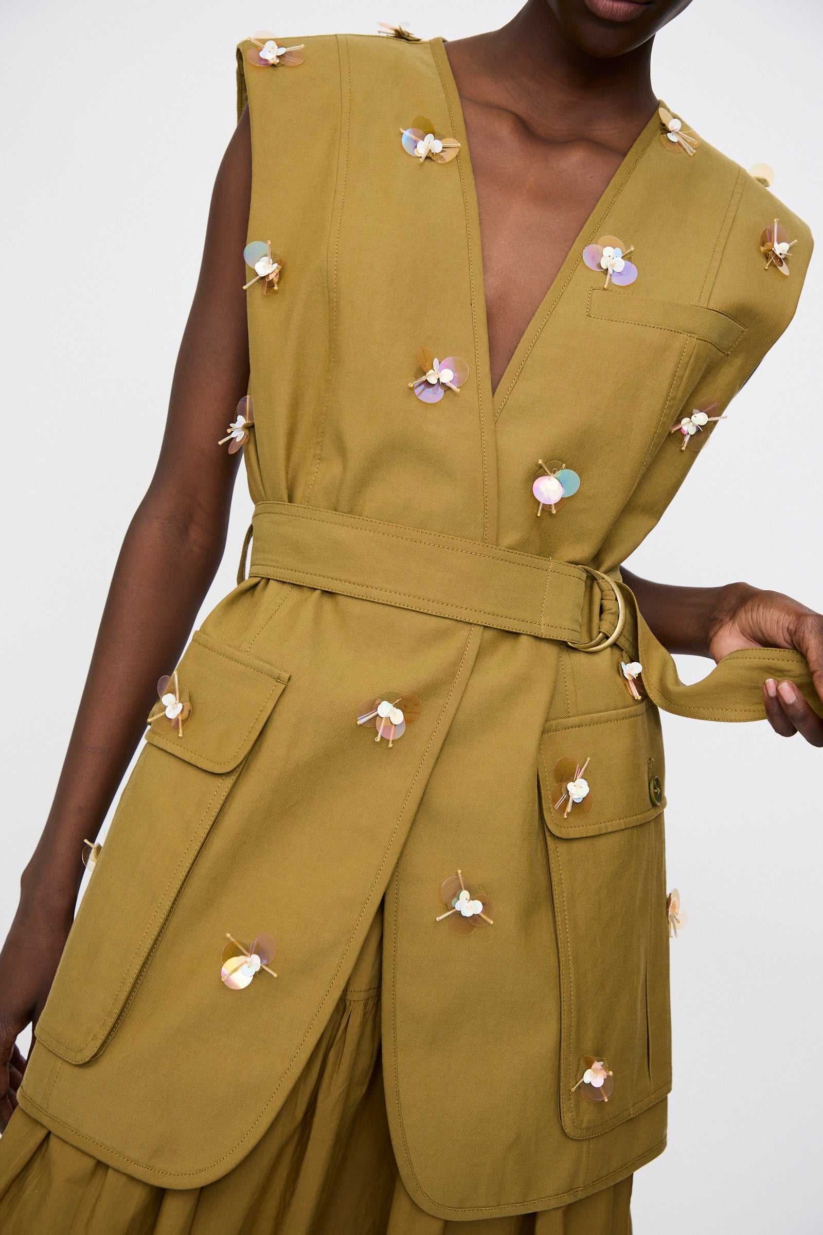 A person wears the Ulla Johnson Embroidered Cotton Twyla Vest in Lichen, a sleeveless, belted vest with large front pockets and small multicolored floral embroidery.