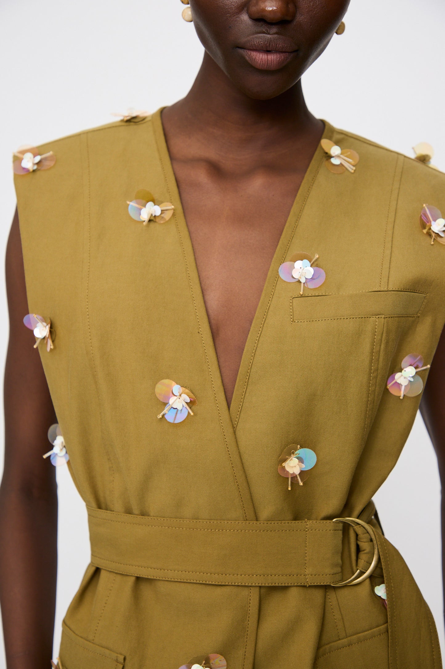 A person wearing the Embroidered Cotton Twyla Vest in Lichen by Ulla Johnson, a sleeveless olive green vest with a belted waist, deep V-neck, and decorative paillette florals, styled like a tailored dress.