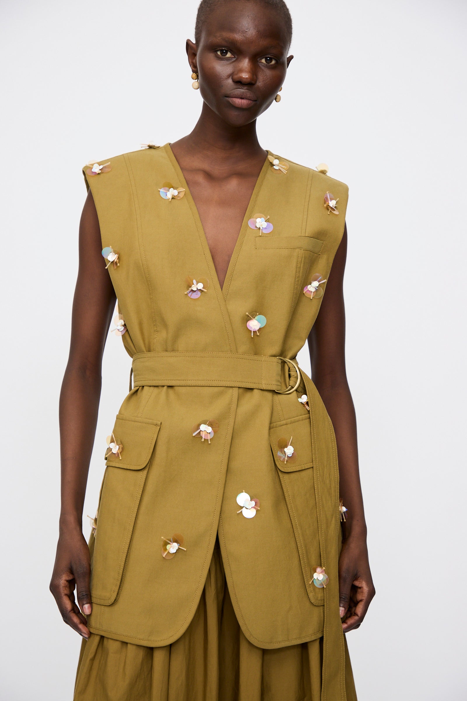 Model wears the Ulla Johnson Embroidered Cotton Twyla Vest in Lichen, a tailored sleeveless vest with large pockets and subtle paillette floral details, against a plain white background.