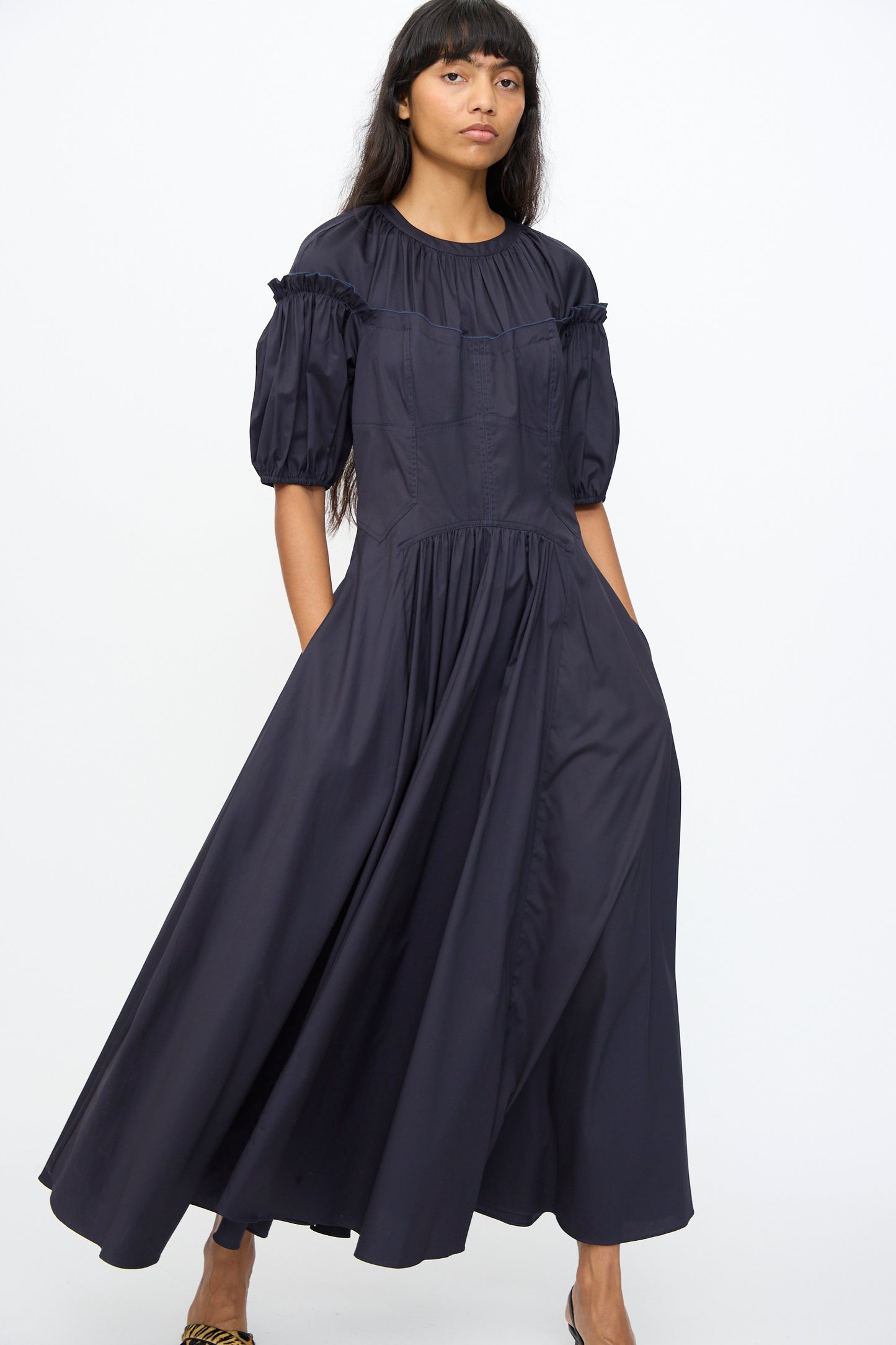 A woman stands against a plain white background wearing the Ulla Johnson Estelle Dress in Midnight, a dark, loose-fitting organic cotton design with puffed short sleeves.