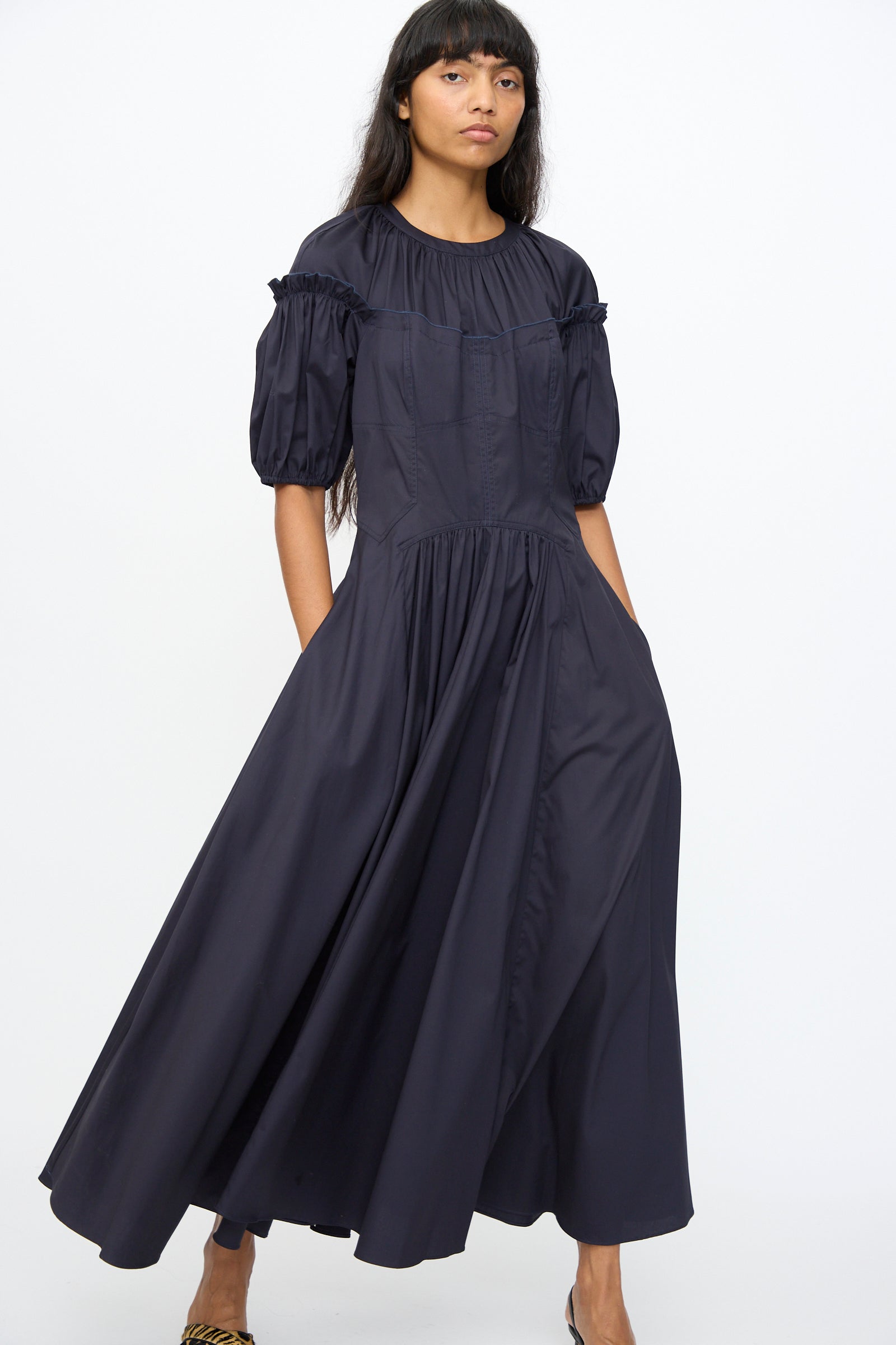 A woman stands against a plain white background wearing the Ulla Johnson Estelle Dress in Midnight, a dark, loose-fitting organic cotton design with puffed short sleeves.