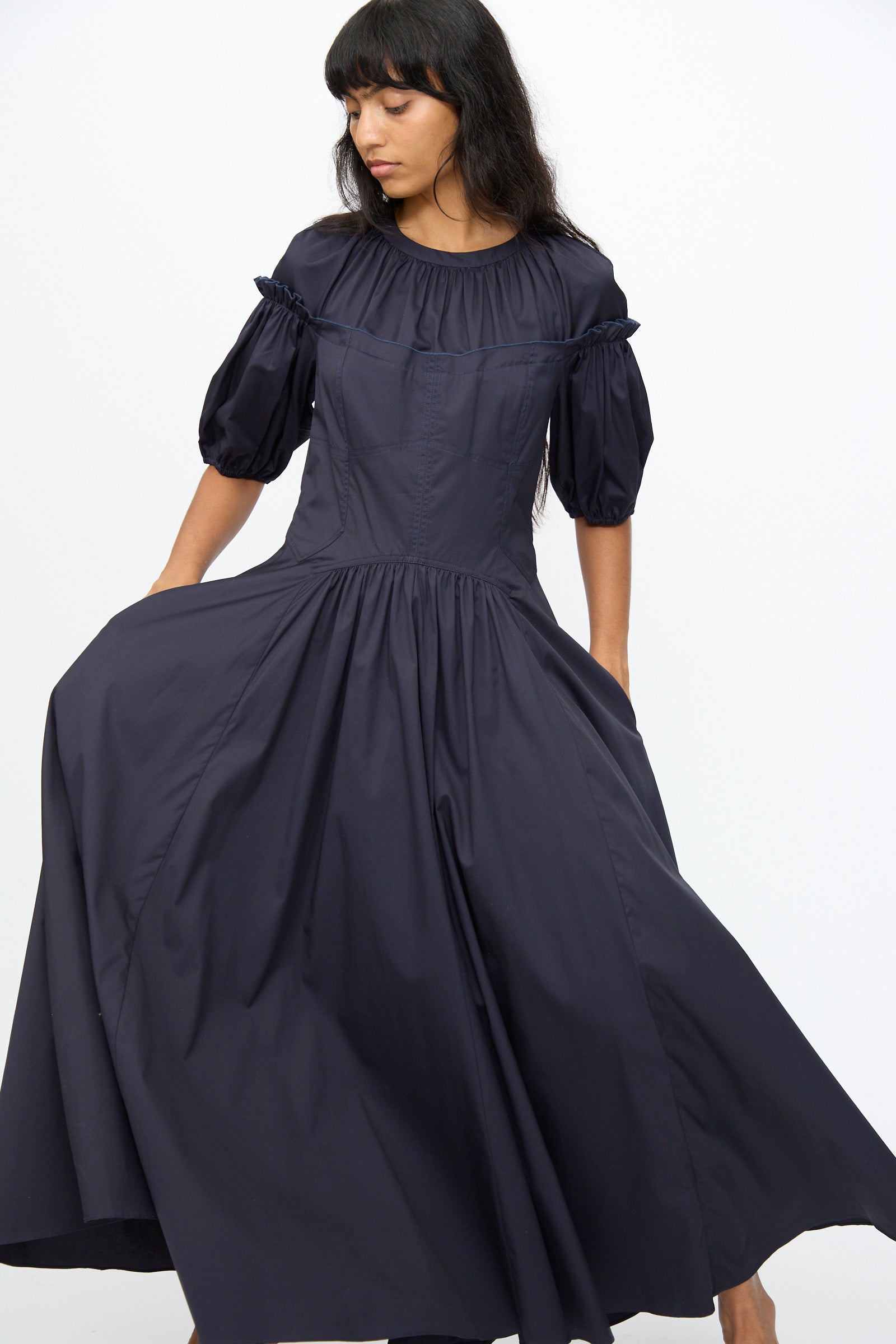 A woman stands against a plain white background, holding out one side of her skirt while wearing the Ulla Johnson Estelle Dress in Midnight, an organic cotton puff sleeve dress with a corset silhouette.