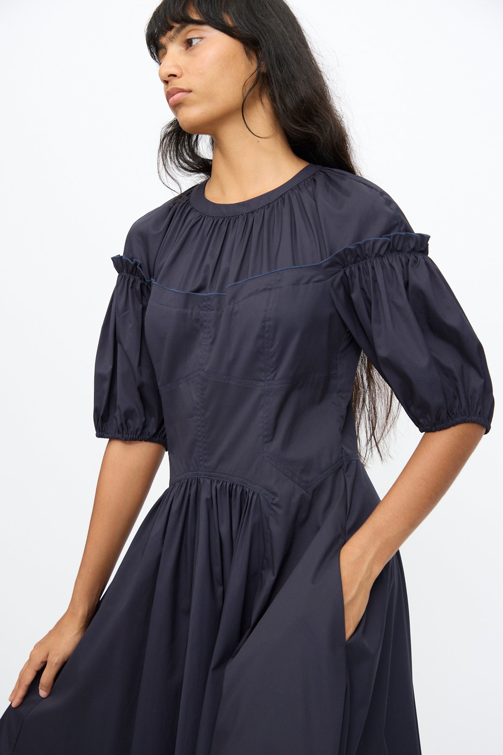 A woman stands against a plain white background wearing the Ulla Johnson Estelle Dress in Midnight, an organic cotton dress featuring puff sleeves and ruffle details.