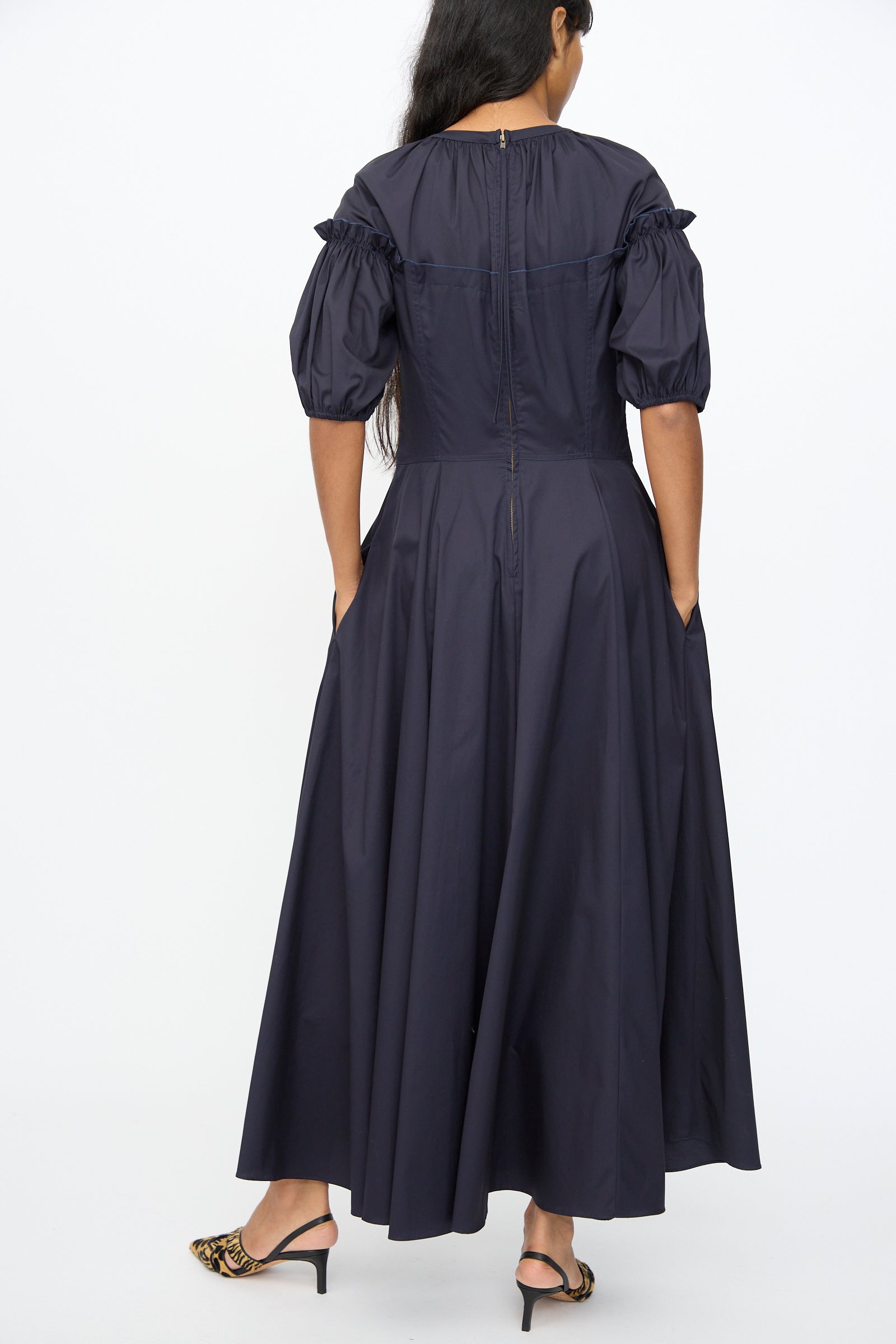 A woman stands facing away, wearing the Ulla Johnson Estelle Dress in Midnight—an organic cotton puff sleeve dress with a fitted waist and pockets—styled with leopard-print heeled sandals.