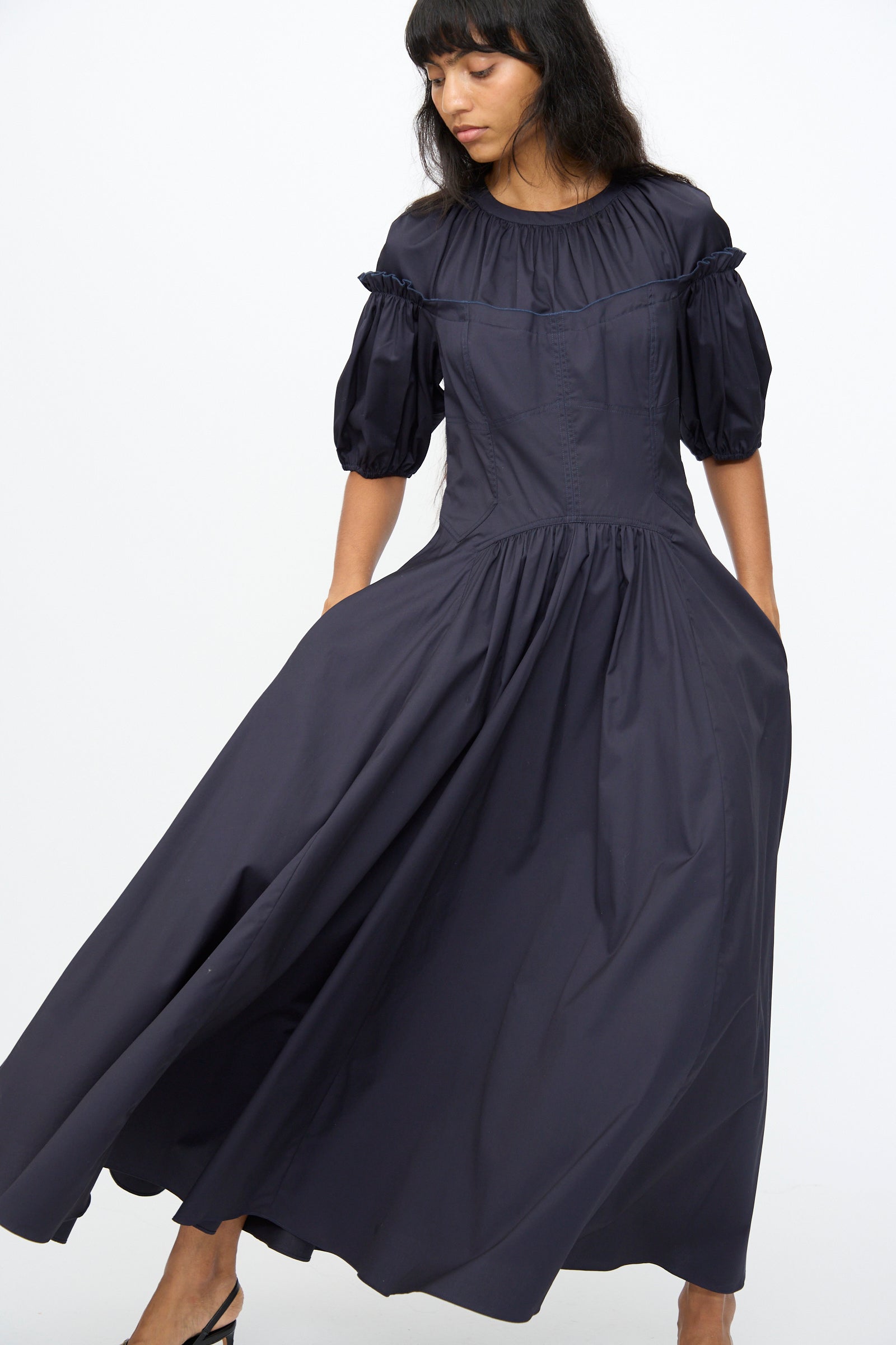 A woman showcases the Ulla Johnson Estelle Dress in Midnight—an organic cotton, long, puff sleeve dress with a gathered neckline and skirt—standing against a plain white background to highlight its timeless elegance.