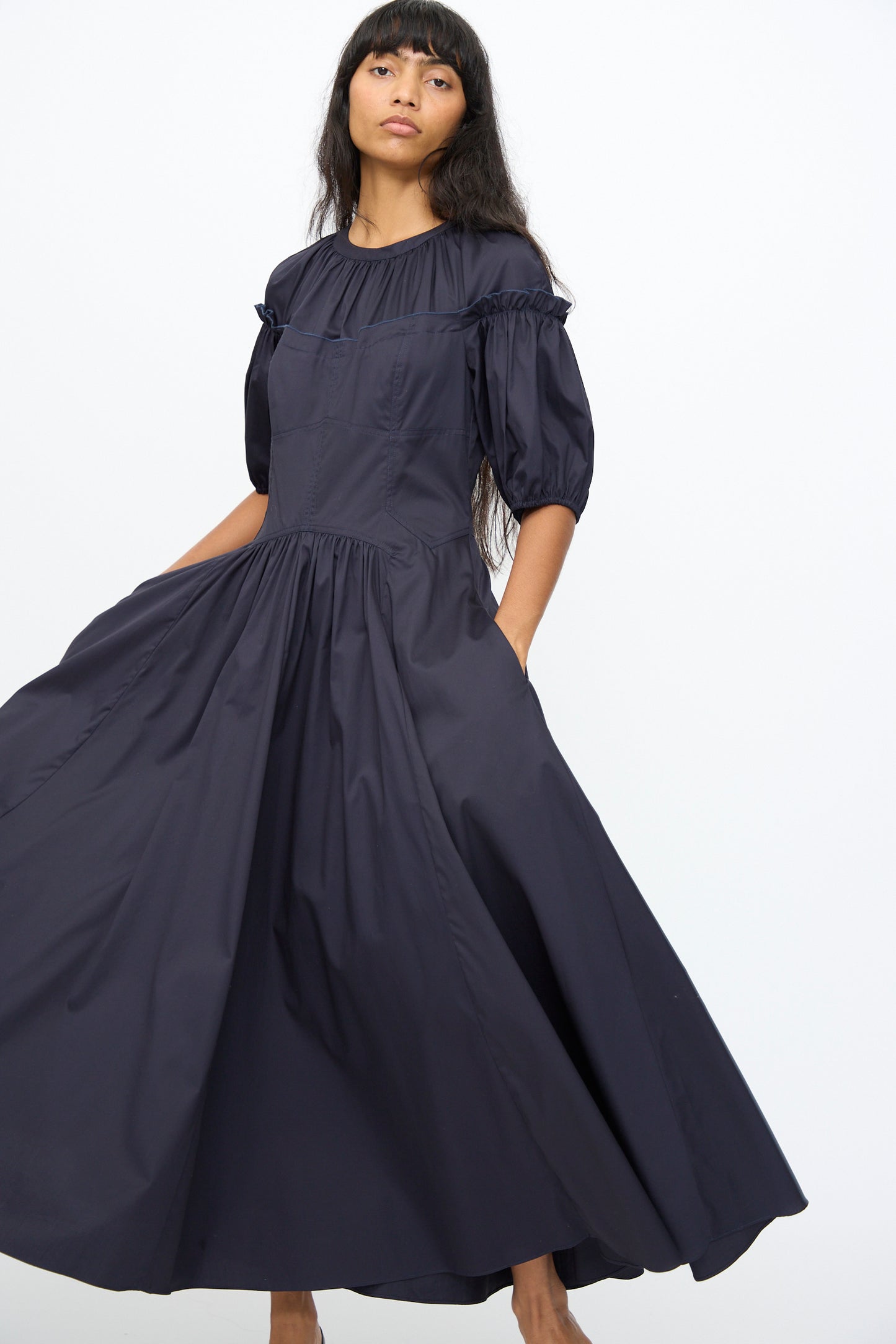 A model wears the Ulla Johnson Estelle Dress in Midnight, a long, dark navy puff-sleeve dress with a gathered waist and ruffle accents that features a subtle corset silhouette, standing against a plain white background.