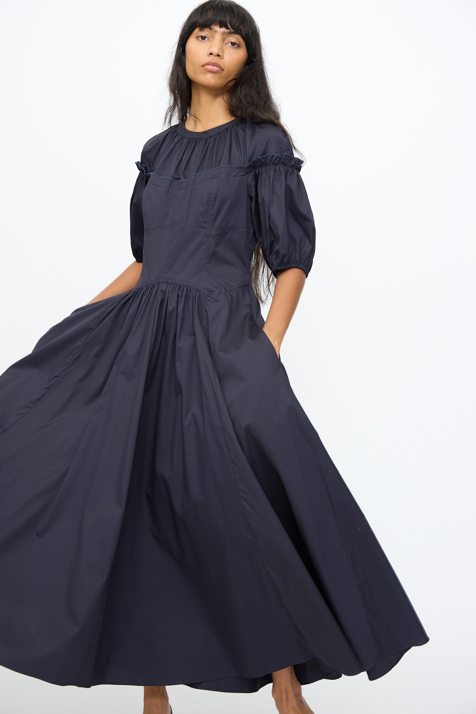 A model wears the Ulla Johnson Estelle Dress in Midnight, a long, dark navy puff-sleeve dress with a gathered waist and ruffle accents that features a subtle corset silhouette, standing against a plain white background.