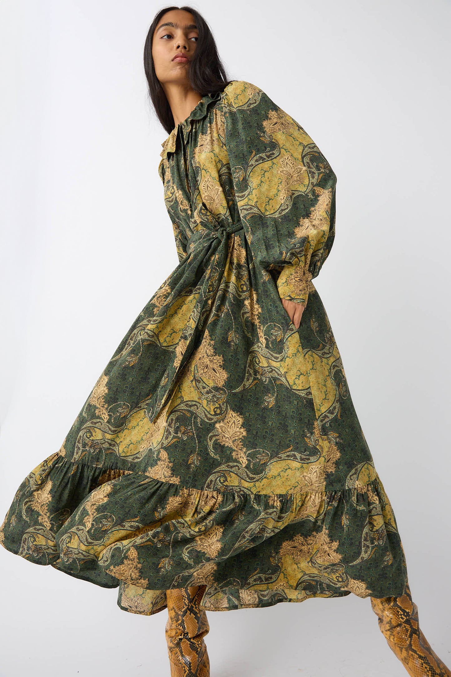 Woman wearing the Fianna Dress in Alpine by Ulla Johnson, a flowing green and yellow paisley print dress with a ruffled neckline, paired with matching yellow boots, posing against a plain light background.