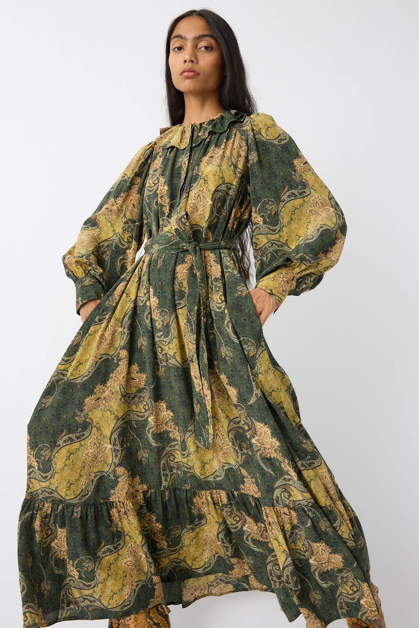 A woman wears the Ulla Johnson Fianna Dress in Alpine, a long-sleeved, ankle-length design featuring a green and gold paisley print. She has long, dark hair and stands against a plain background.