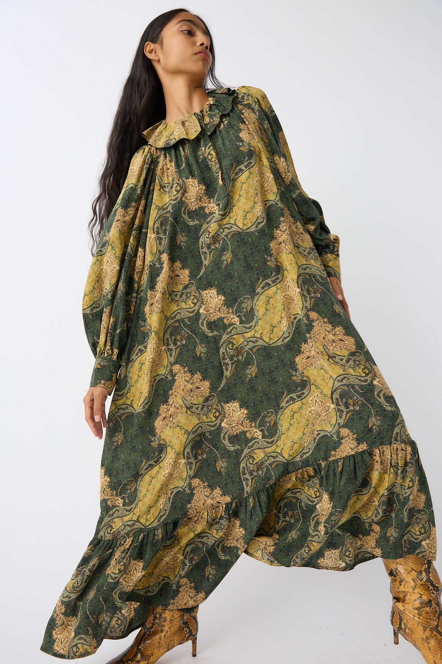 A person with long hair wearing an oversized Ulla Johnson Fianna Dress in Alpine, featuring a green and yellow paisley print, poses against a plain background.