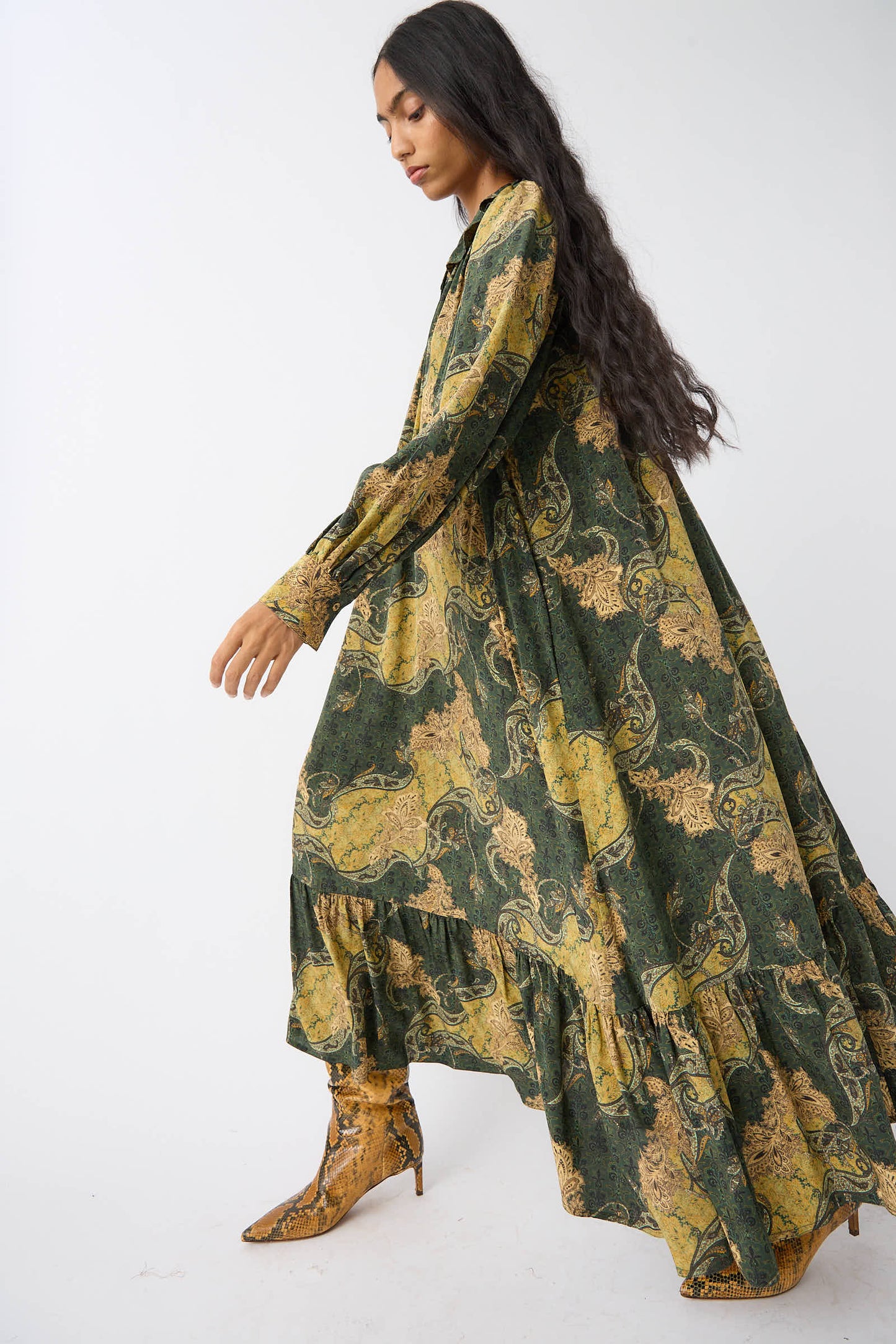 A person with long hair wearing a green and yellow Ulla Johnson Fianna Dress in Alpine and snakeskin-patterned boots walks against a plain white background.