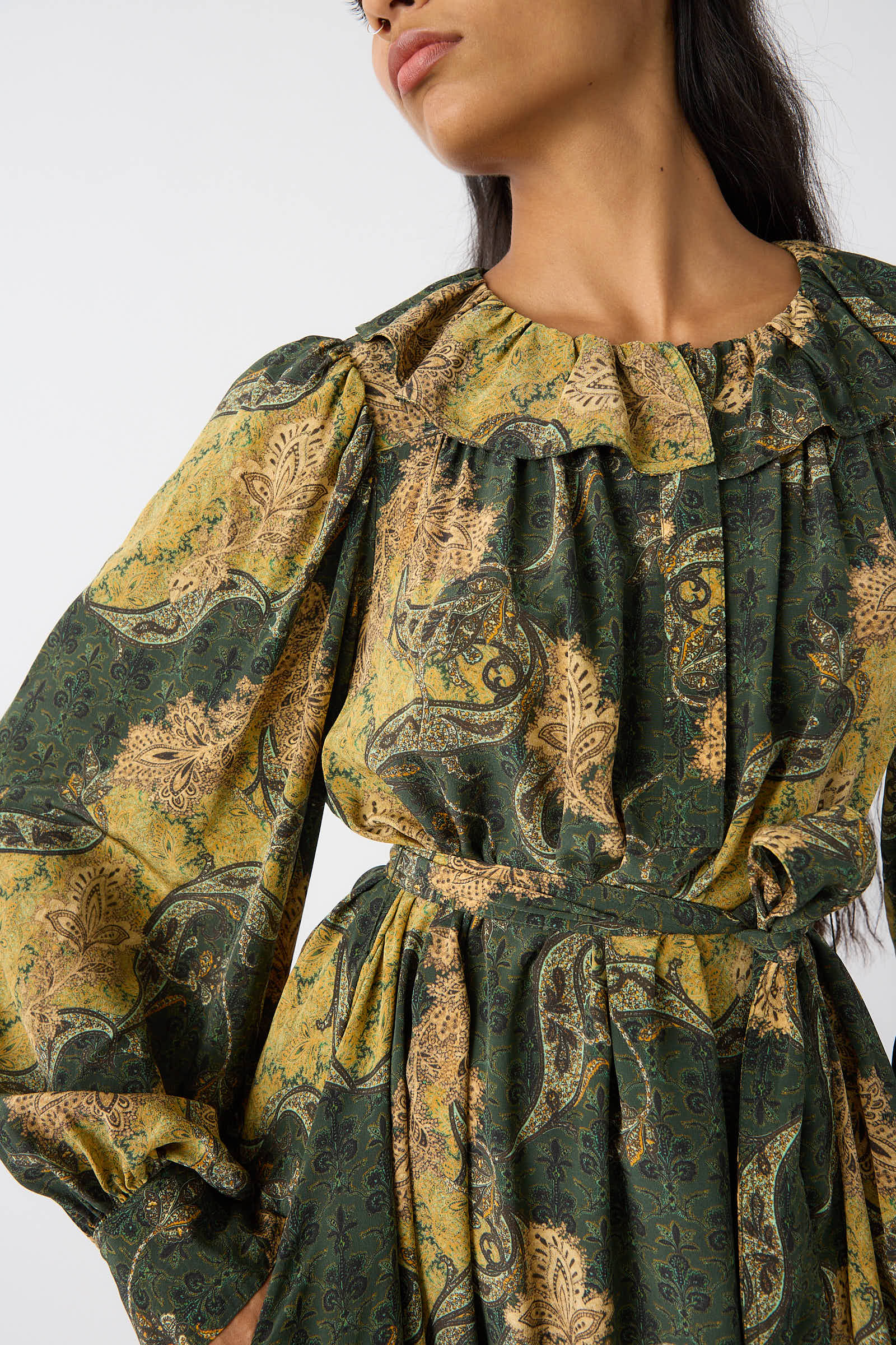 A person with long hair wears the Fianna Dress in Alpine by Ulla Johnson, which features a green and gold paisley print, puffed sleeves, and a high ruffled neckline.