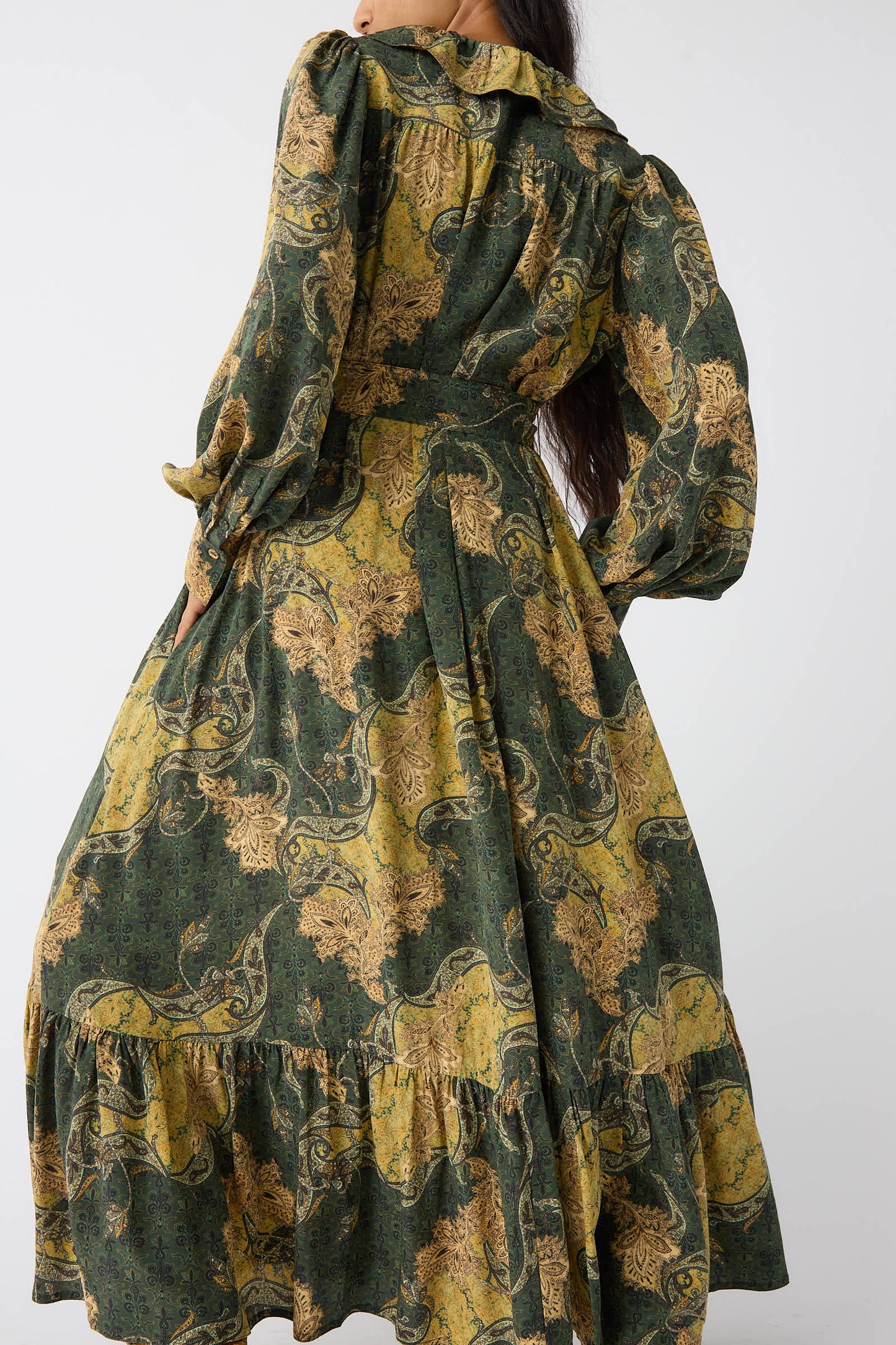 A person wearing the Fianna Dress in Alpine by Ulla Johnson, a long-sleeved, paisley print green and yellow dress with ruffles, is shown from the back.