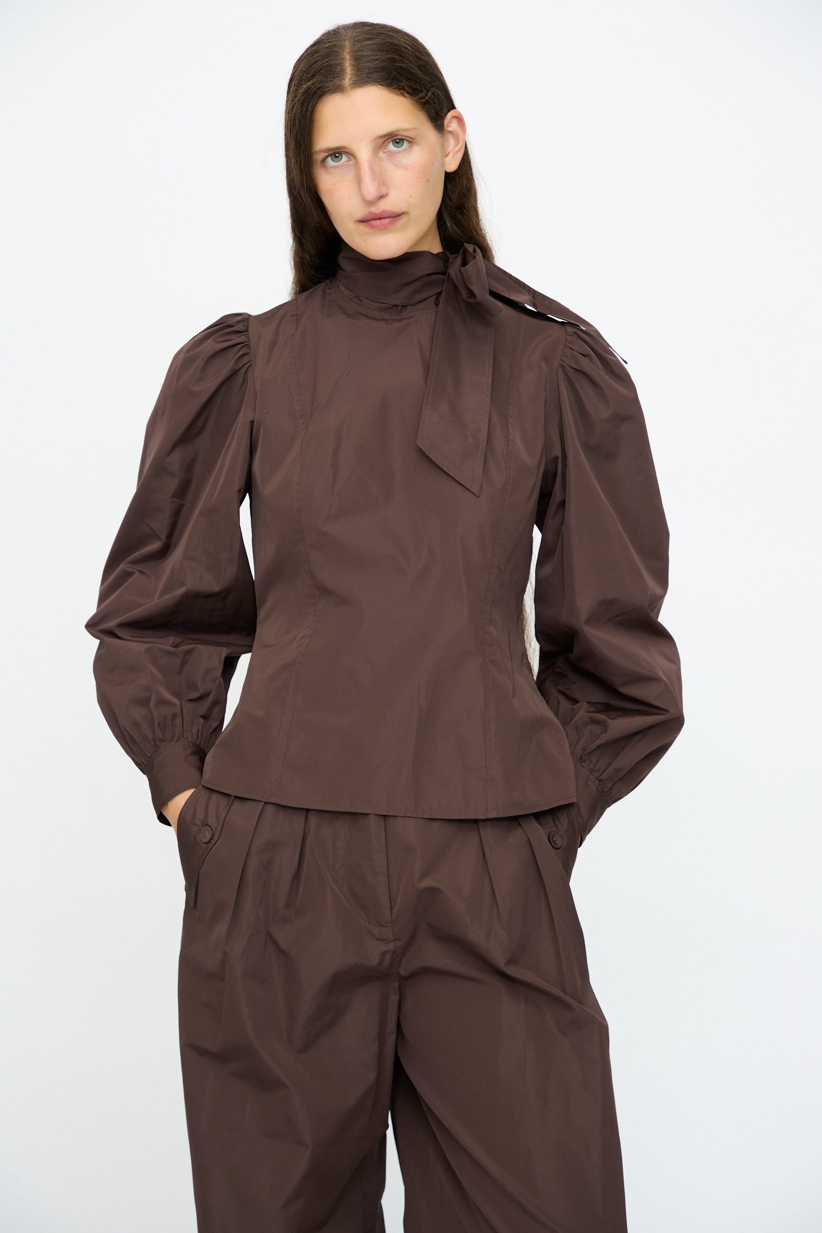 A woman wears the Ulla Johnson Giorgia Silk Blouse in Sequoia, featuring balloon sleeves and a bow neck, paired with matching high-waisted, wide-leg pants, standing against a plain white background.