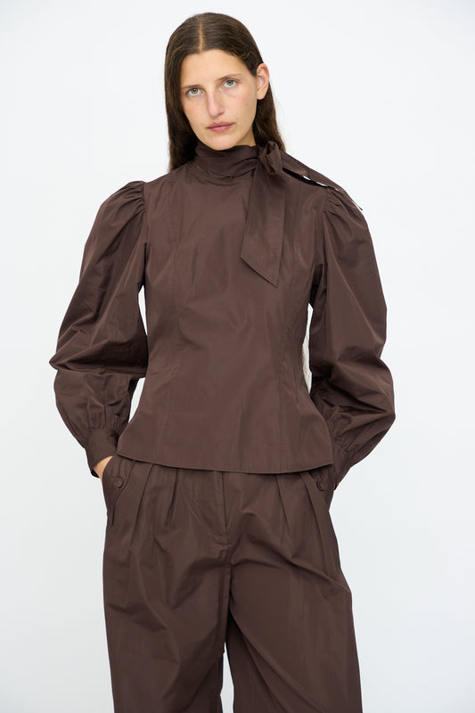 A woman wears the Ulla Johnson Giorgia Silk Blouse in Sequoia, featuring balloon sleeves and a bow neck, paired with matching high-waisted, wide-leg pants, standing against a plain white background.