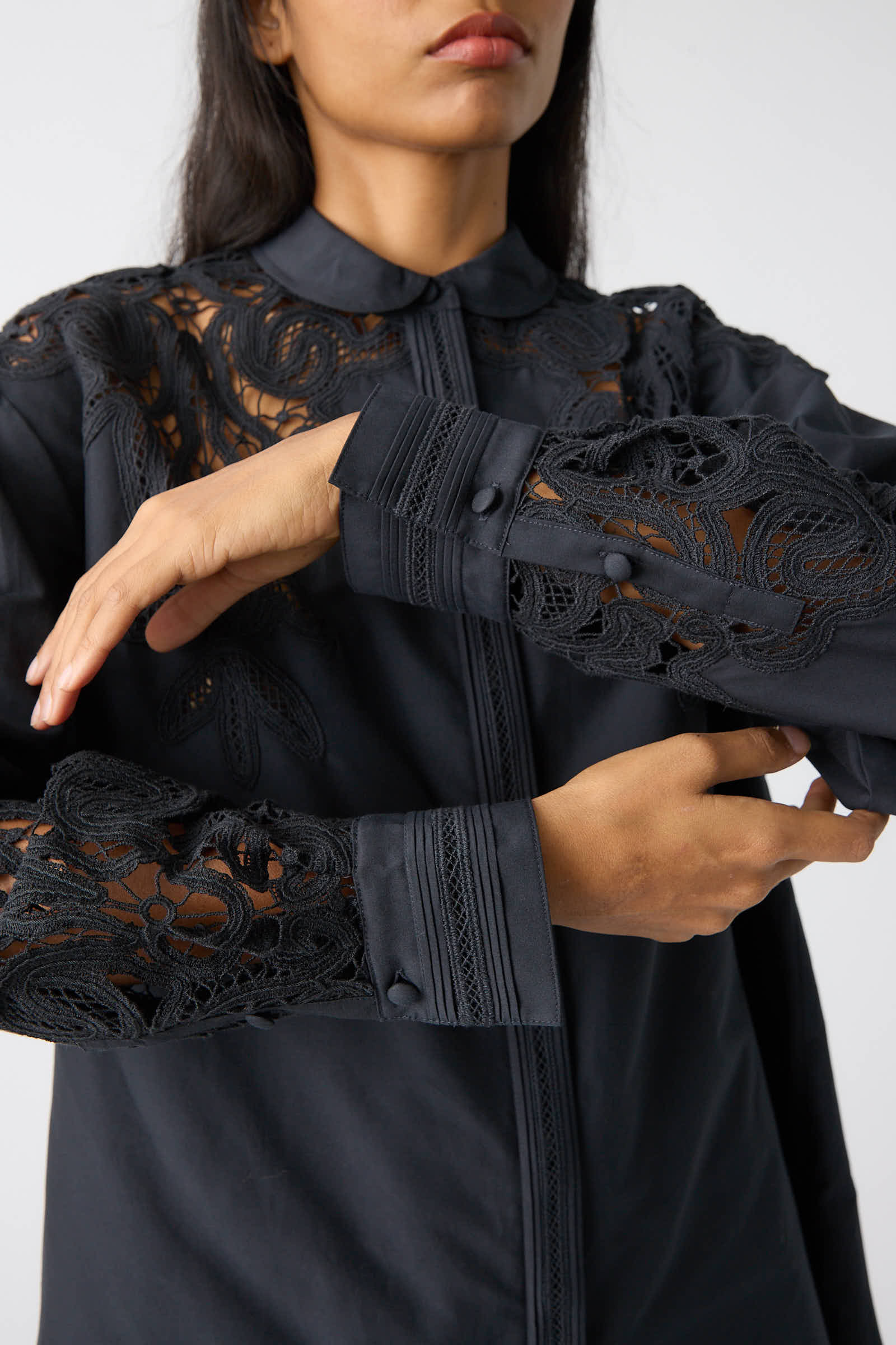 A person wearing the Ulla Johnson Harden Tunic in Noir, featuring intricate Battenberg lace detailing on the shoulders and long sleeves.