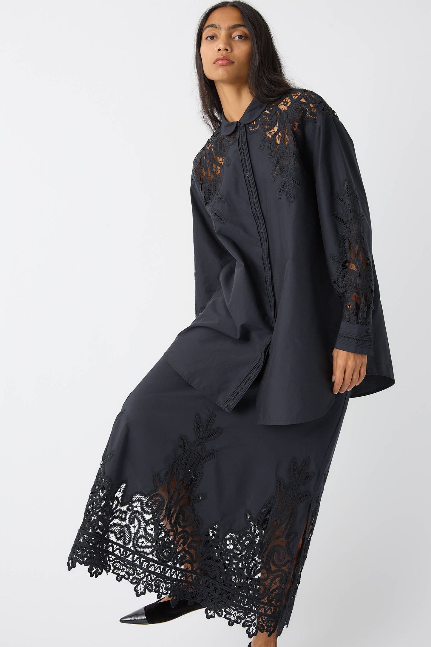 A person wearing the Ulla Johnson Harden Tunic in Noir, a long-sleeve dress made from a cotton/silk blend with Battenberg lace details on the shoulders, sleeves, and hemline, standing against a plain white background.