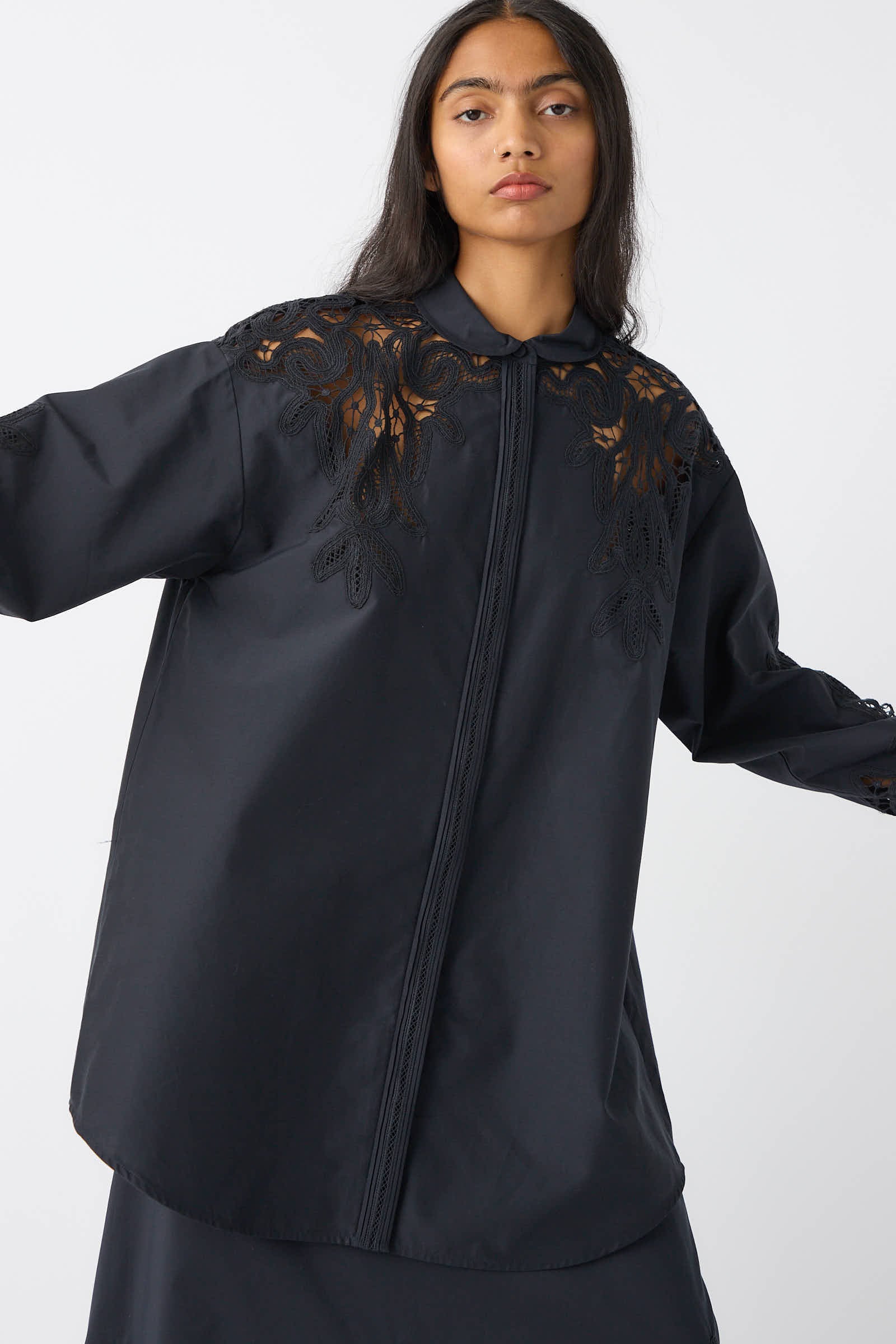 A person wearing the Ulla Johnson Harden Tunic in Noir, featuring intricate Battenberg lace detailing on the shoulders. This oversized tunic with long sleeves and a loose fit is made from a cotton/silk blend.