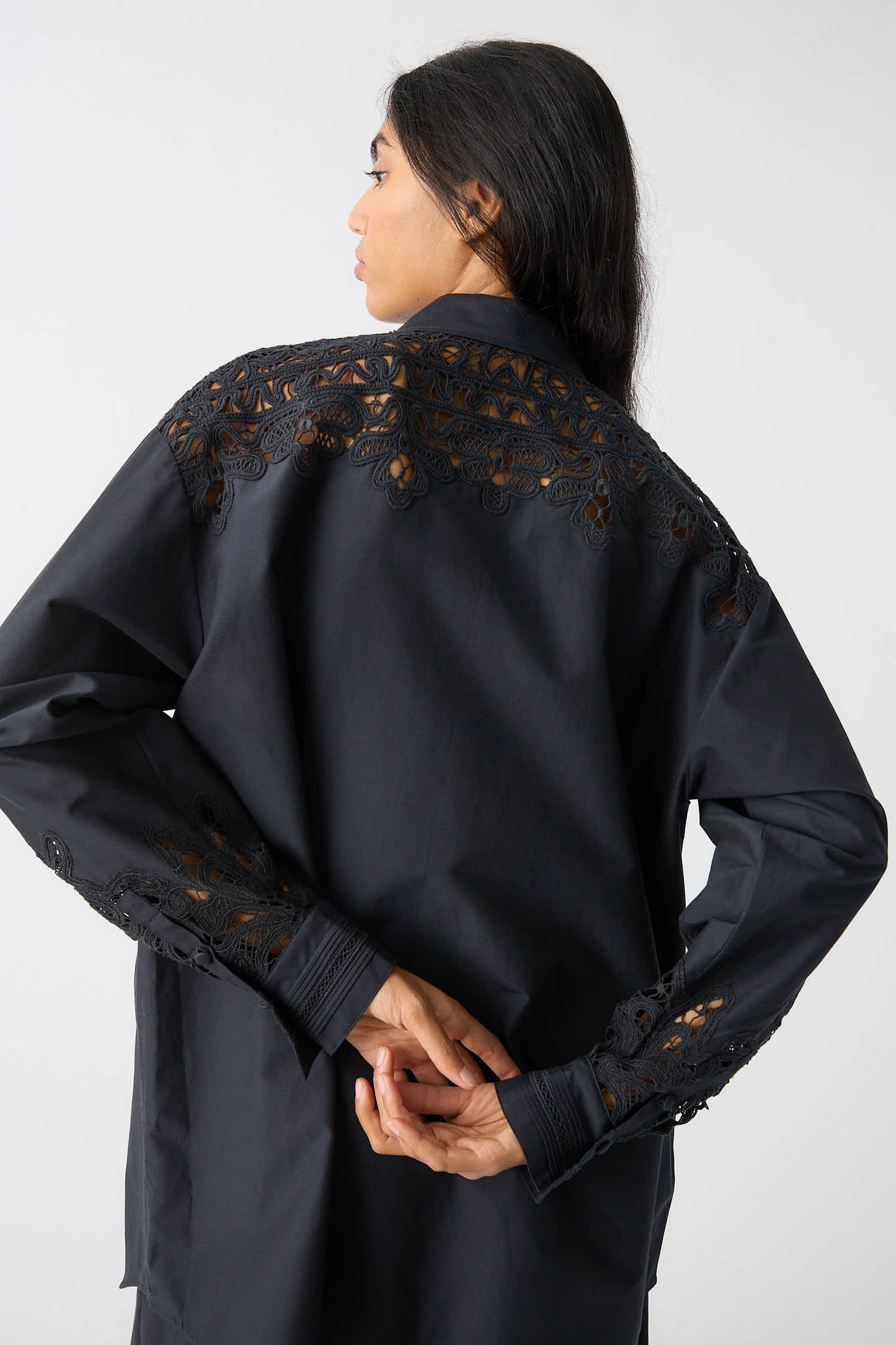 A person with long dark hair facing away, wearing the Ulla Johnson Harden Tunic in Noir, featuring intricate Battenberg lace details on the back and sleeves, and hands clasped behind their back.